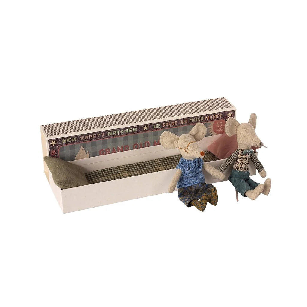 Two small Maileg Mice from Maileg, dressed in clothes, sit by an open matchbox-style bed with pillow and blanket. The box features vintage designs, making it a charming addition to your mouse family collection.