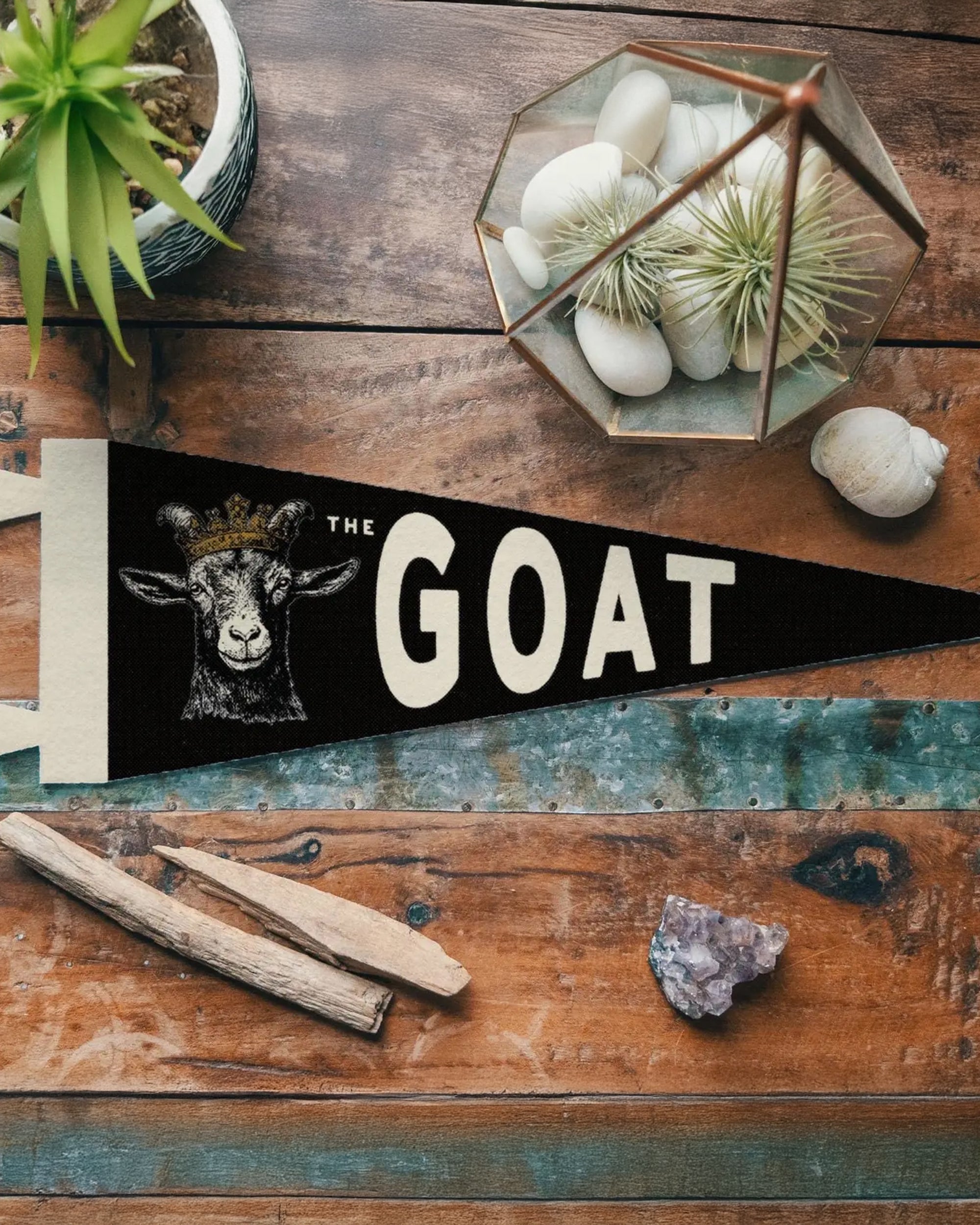 The GOAT Felt Pennant by Faire, in black vintage felt and featuring a crowned goat design, sits on a rustic wooden table with a plant, an amethyst crystal, and a glass terrarium filled with white stones.