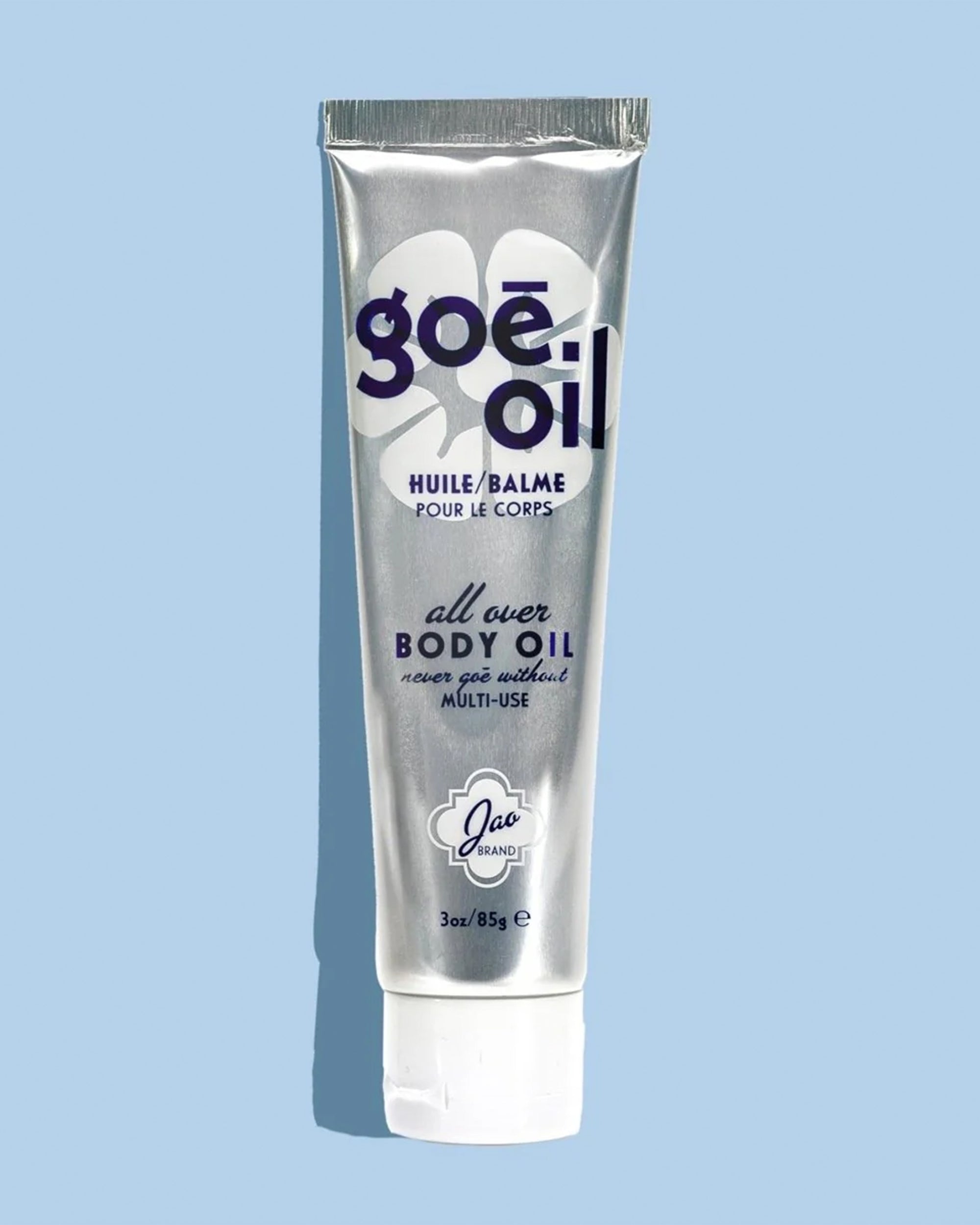 A silver tube of Goe Oil by Jao Limited with purple and black text and a white cap stands upright on a light blue background, featuring a white flower design behind it. Perfect for moisturizing and hydrating skin.
