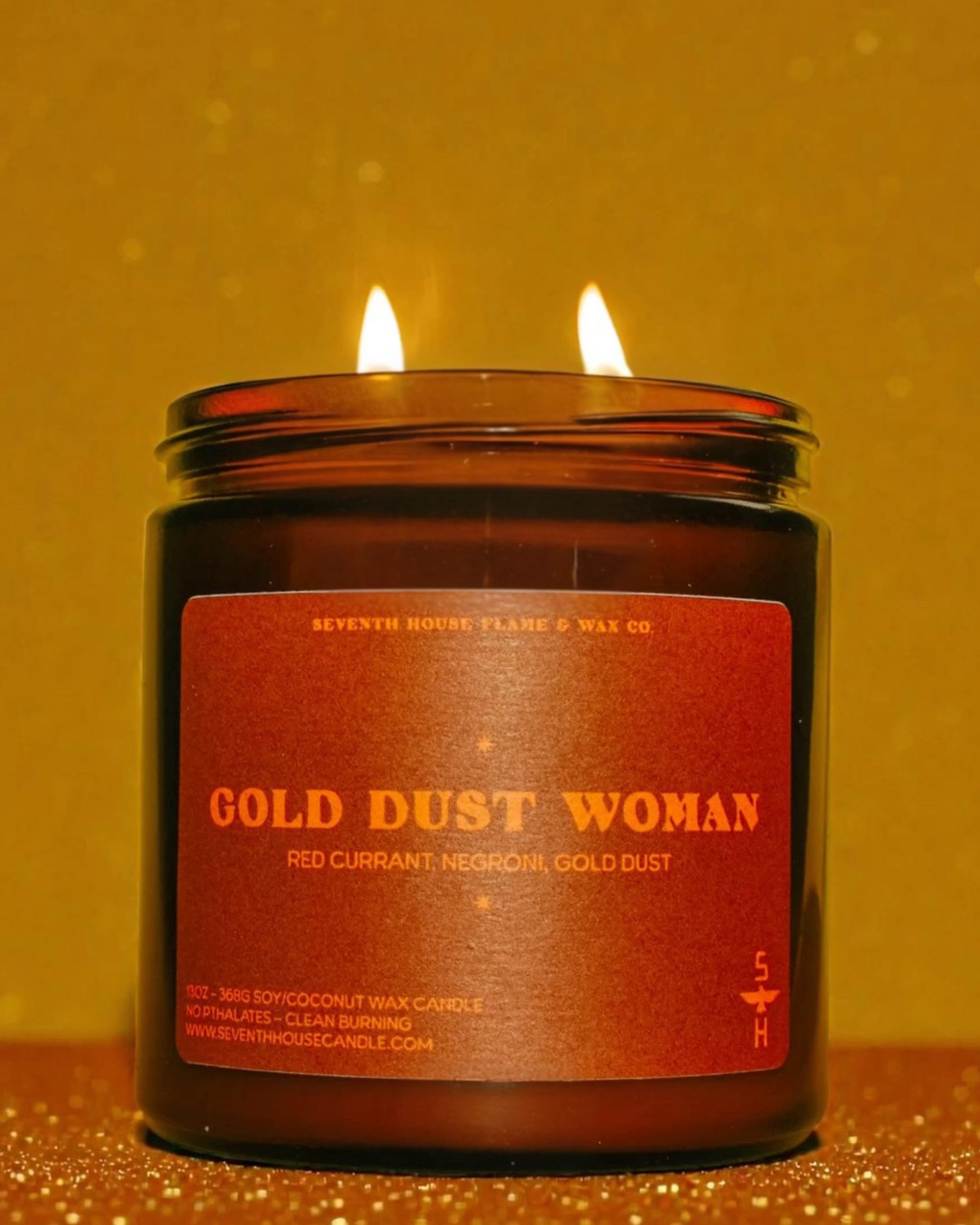Gold Dust Woman Candle by Faire with two glowing flames, set against a shimmering gold backdrop.