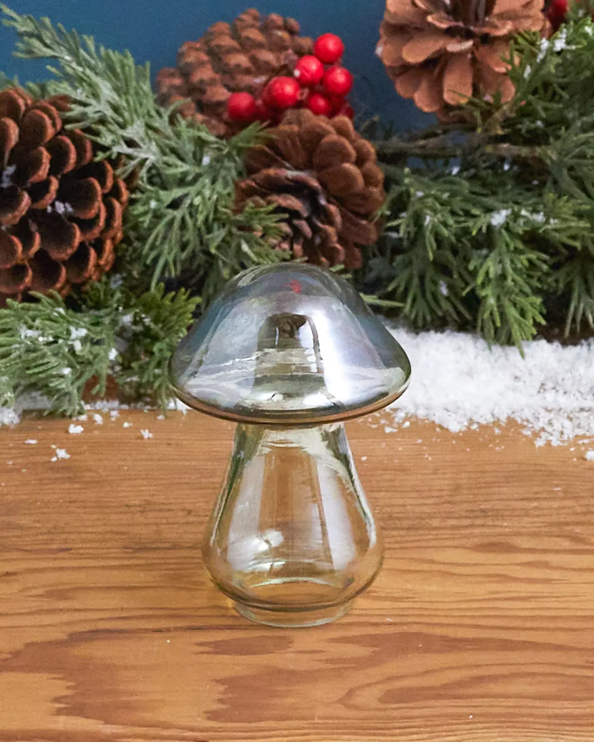 The Be Home Gold Luster Glass Mushroom S rests on a wooden surface, surrounded by pinecones, red berries, evergreen branches, and artificial snow, creating a festive winter scene.