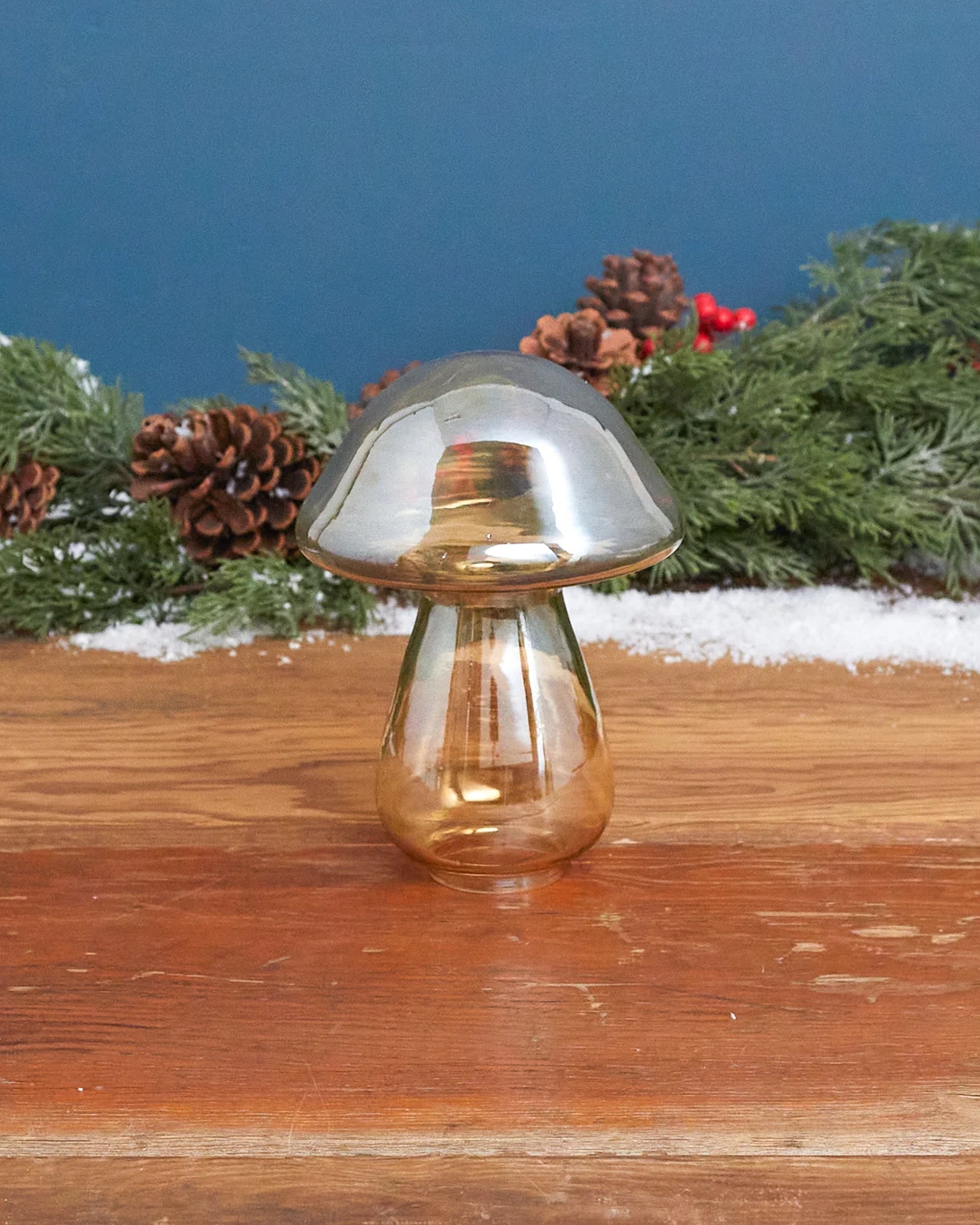 The Be Home Gold Luster Glass Mushroom L rests on wood, framed by pine branches, pine cones, red berries, and artificial snow for a festive display.