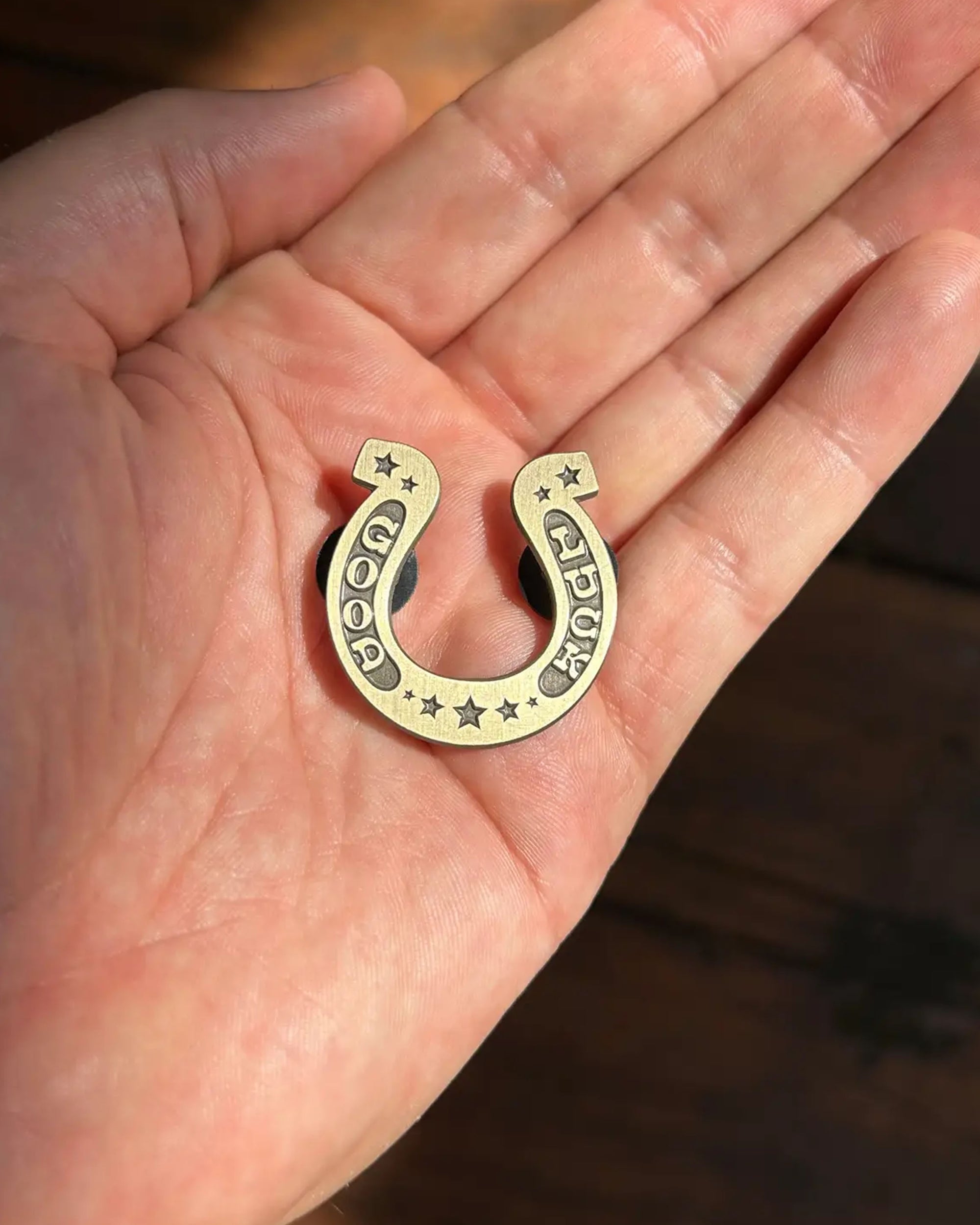 A hand holds Faire’s Good Luck Pin, a small gold-colored charm engraved with stars and “GOOD LUCK.” Sunlight casts soft shadows on the palm and wooden surface below.