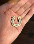 A hand holds Faire’s Good Luck Pin, a small gold-colored charm engraved with stars and “GOOD LUCK.” Sunlight casts soft shadows on the palm and wooden surface below.