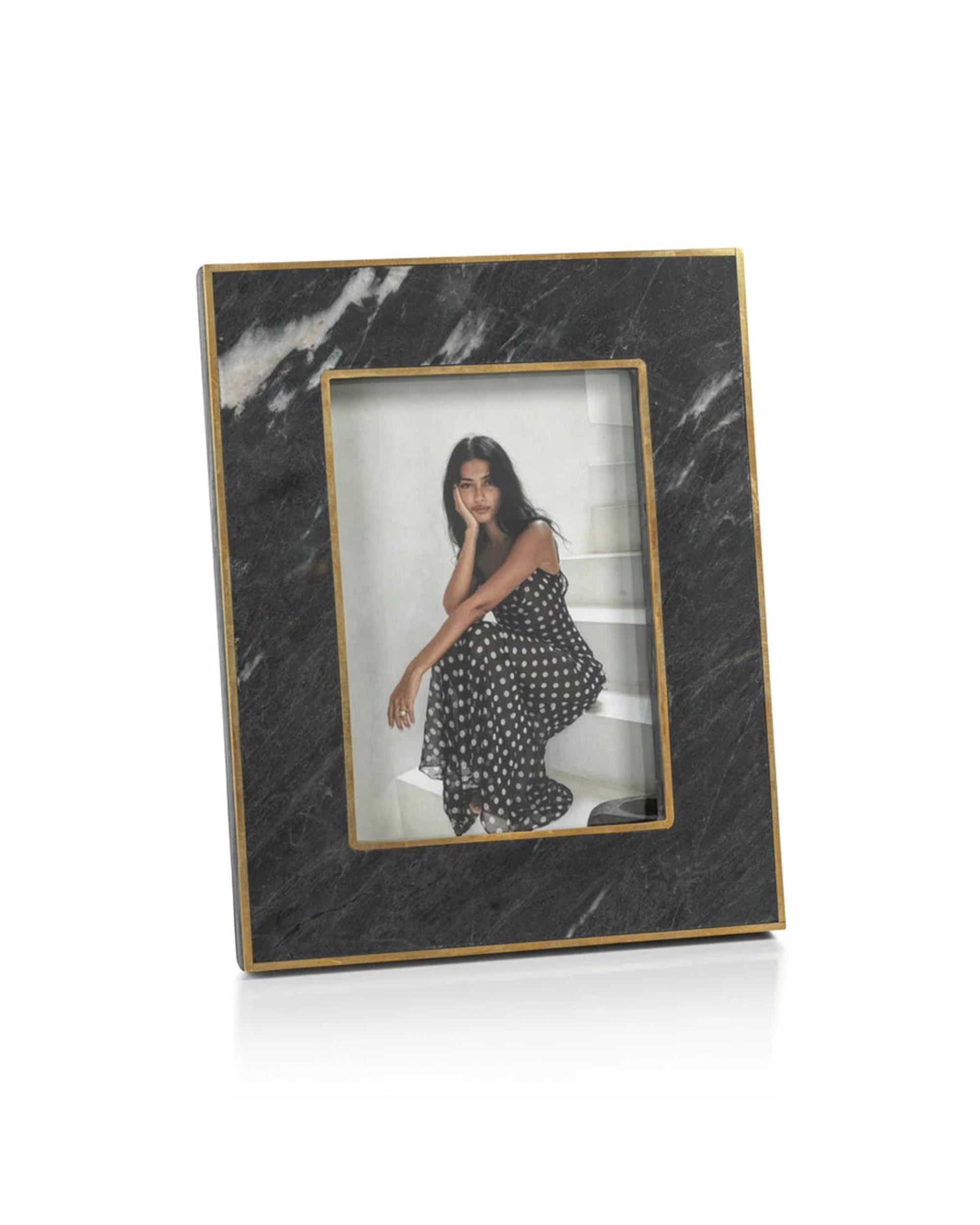 The Zodax IN-7597 Granite Photo Frame 5x7 showcases a photo of a woman in a black and white polka dot dress, seated with her chin on her hand. This elegant frame stands gracefully on a white surface.