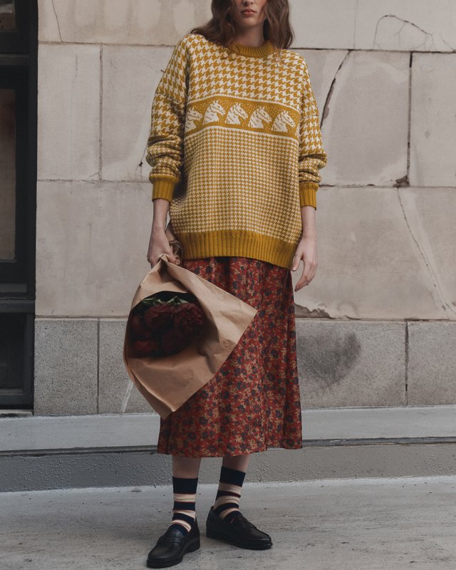 Model wearing a mustard patterned sweater with a red floral midi skirt, striped socks and dark shoes, holding a bouquet.