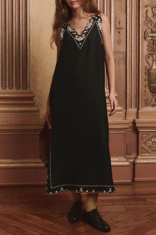 Model in a long black slip dress with pale floral embroidery at the neckline and hem, worn with yellow socks and dark shoes.
