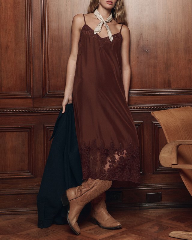 Model in a chocolate slip dress, cream neck bandana and tan cowboy boots, holding a dark shirt beside wood paneling.