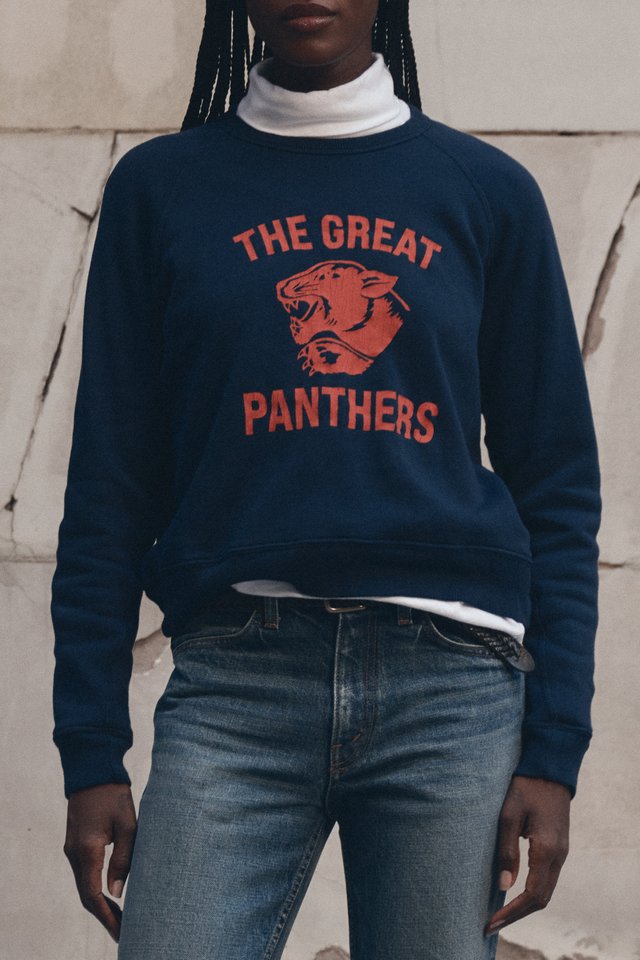 Model in a navy THE GREAT. PANTHERS sweatshirt layered over a white high neck with faded blue jeans.