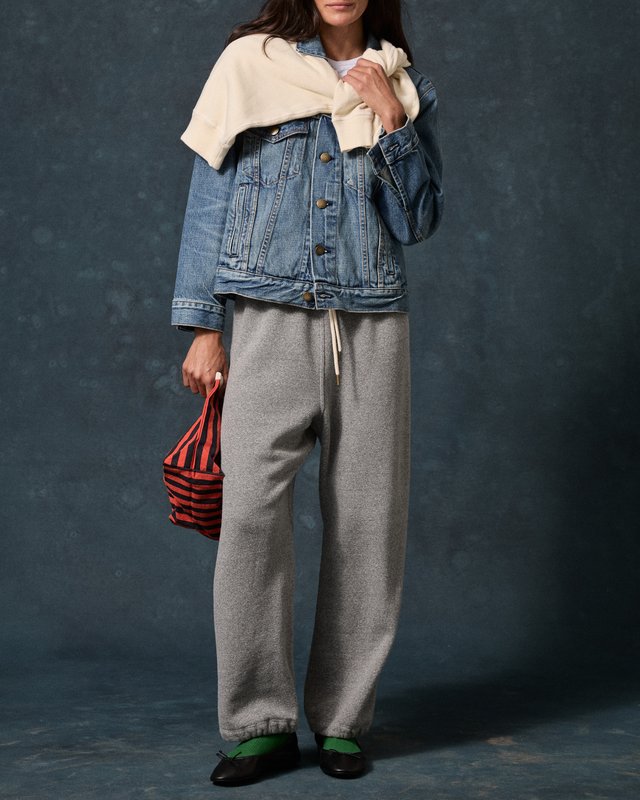 Model in a denim jacket and loose grey sweatpants, with a cream sweater draped over the shoulders and green shoes.