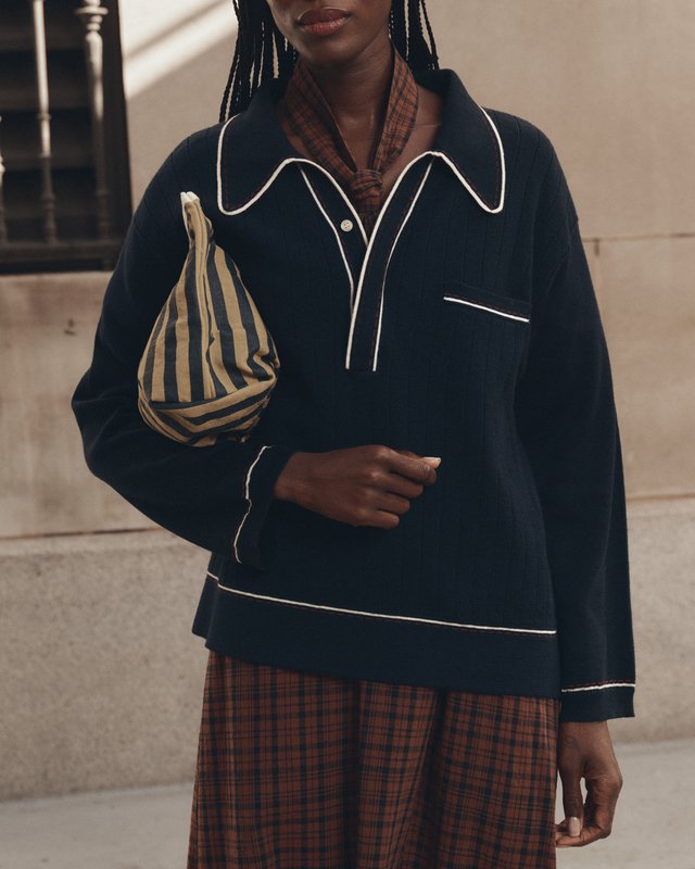 Model wearing a navy top with cream piping over a brown plaid dress and carrying a small striped bag.