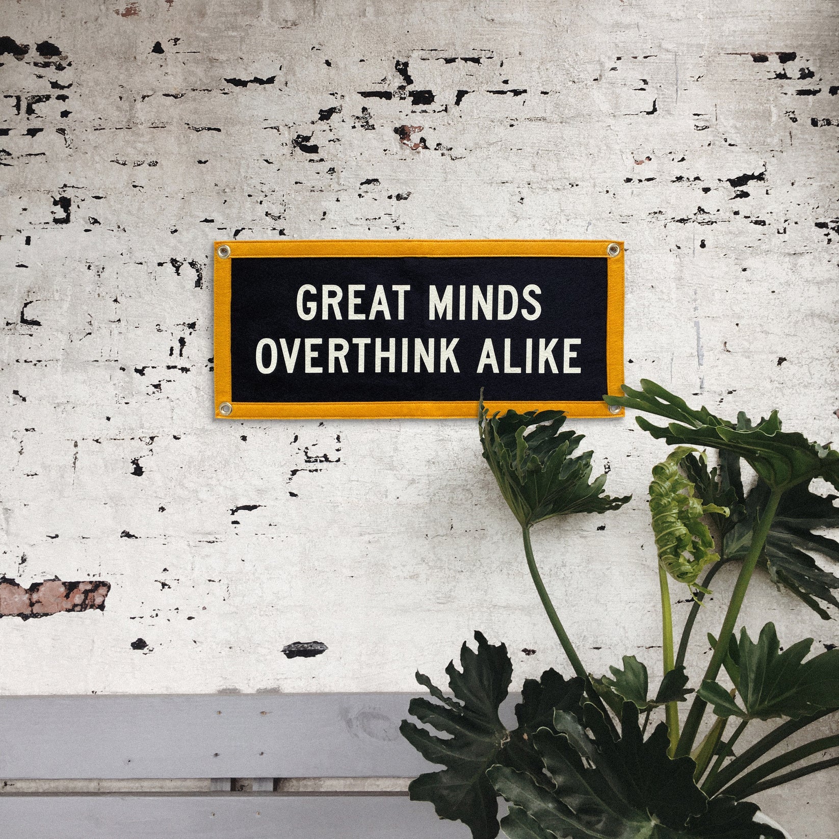 The Oxford Pennant "Great Minds Overthink Alike 10x24" camp flag, featuring a yellow border, hangs on a white brick wall with large green leaves partially obscuring its lower right corner.