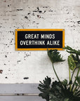 The Oxford Pennant "Great Minds Overthink Alike 10x24" camp flag, featuring a yellow border, hangs on a white brick wall with large green leaves partially obscuring its lower right corner.