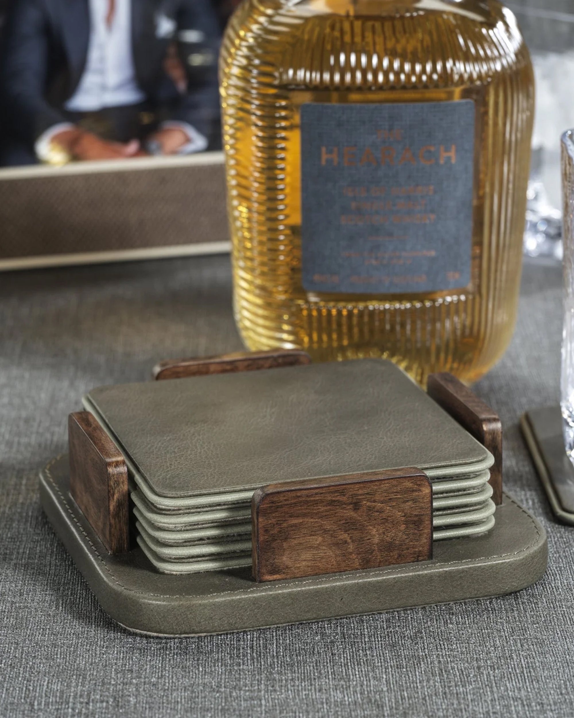 A set of Zodax IN-8237 Coaster S/6 with wooden holders rests on a gray surface beside a glass and a "Hearach" whisky bottle, while a blurred figure appears in the background.