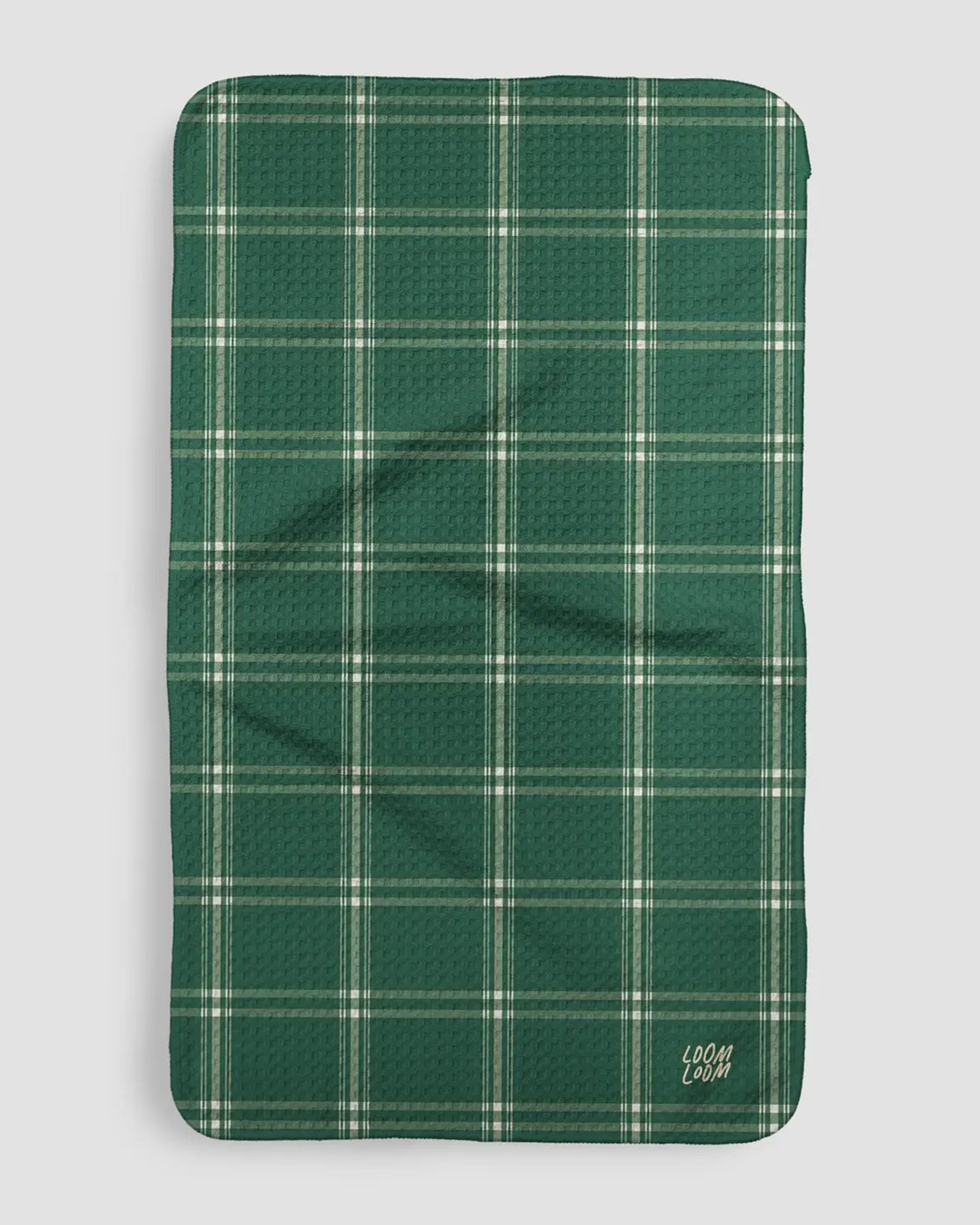 The Faire Evergreen Plaid Towel is a rectangular green and white plaid mat made from EcoLoom fabric, featuring "LOOM & LOOM" in white script in one corner. It is shown laid flat on a plain light background.