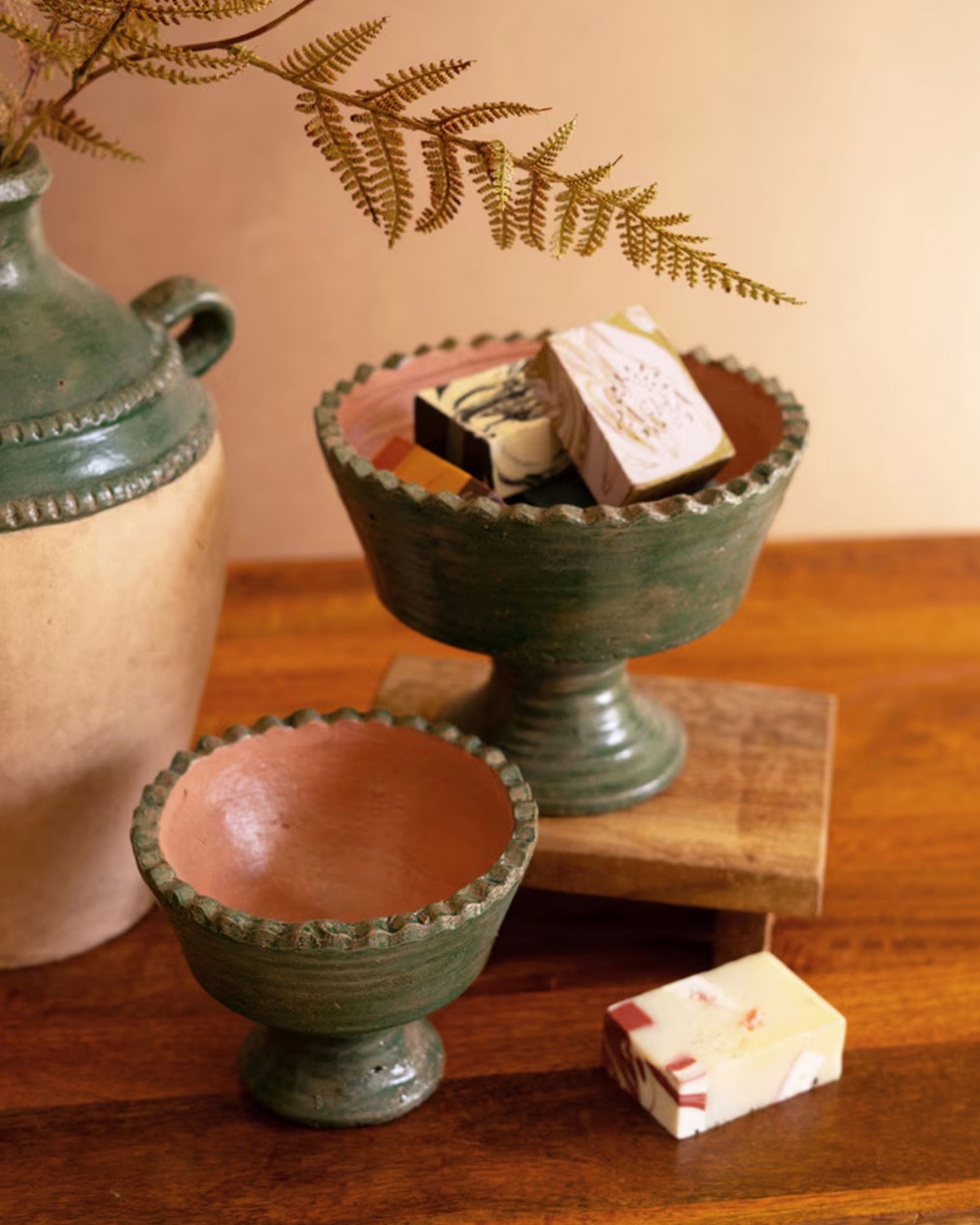 Two Green Terra Clay Compote bowls by Kalalou, Inc sit on a wooden surface—one holds decorative soap bars. A beige vase with ferns stands nearby, and a soap bar rests in front of the bowls.