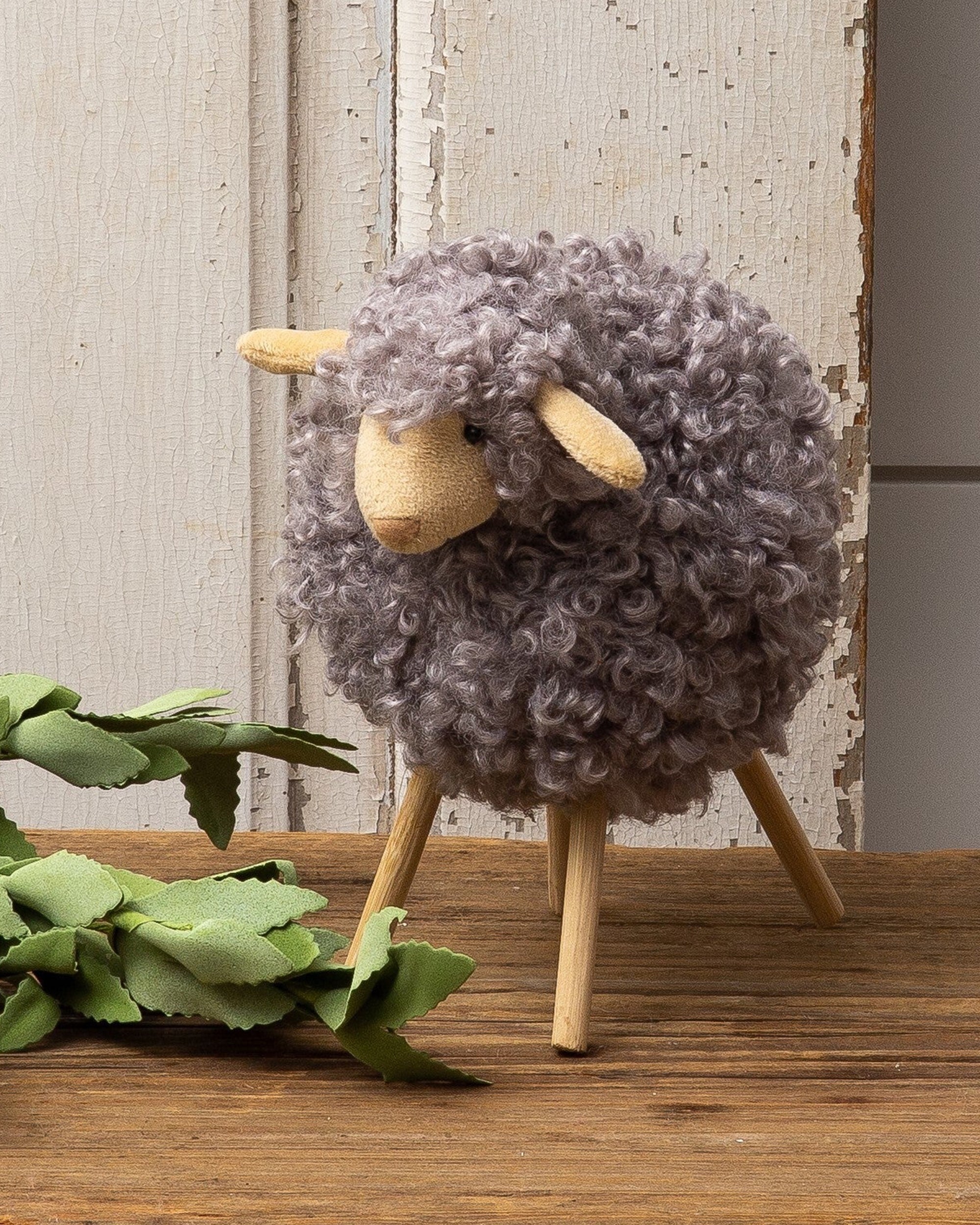 The Gray Sheep Wood Legs figurine by Faire, featuring curly grey wool and wooden stick legs, adds charming decor to any space—displayed on a wooden surface with green fabric leaves against a rustic white background.