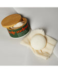 A round white soap bar with a natural scent rests on fabric next to an open container labeled “Faire Grounding Stone Body Lotion,” evoking luxury and suggesting its moisturizing benefits.