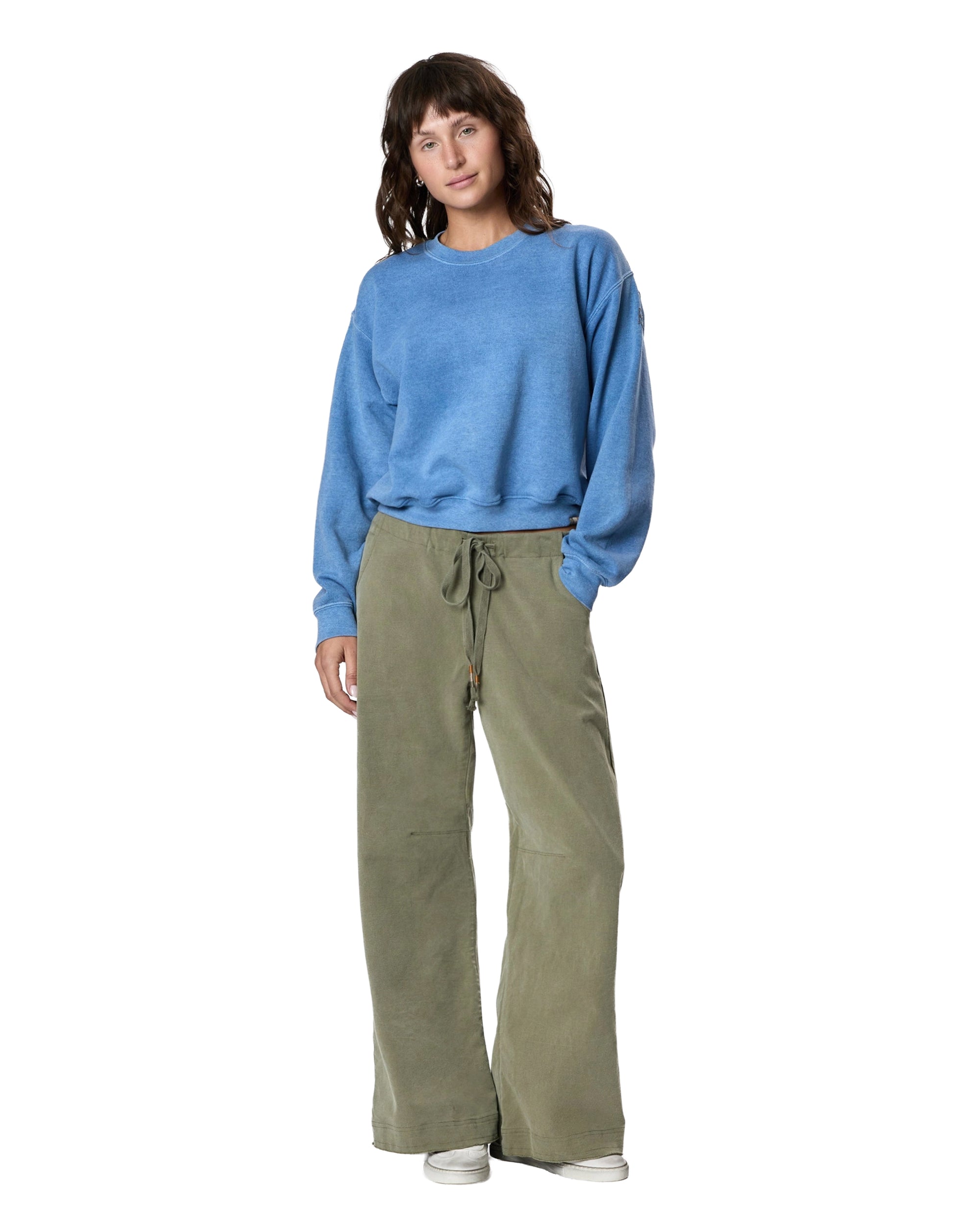 A person with wavy brown hair poses casually against a white background, wearing a loose blue sweatshirt, Faire’s Gwen Boyfriend Wide Leg Pant in olive green organic cotton with a drawstring waist, and white sneakers.