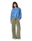 A person with wavy brown hair poses casually against a white background, wearing a loose blue sweatshirt, Faire’s Gwen Boyfriend Wide Leg Pant in olive green organic cotton with a drawstring waist, and white sneakers.