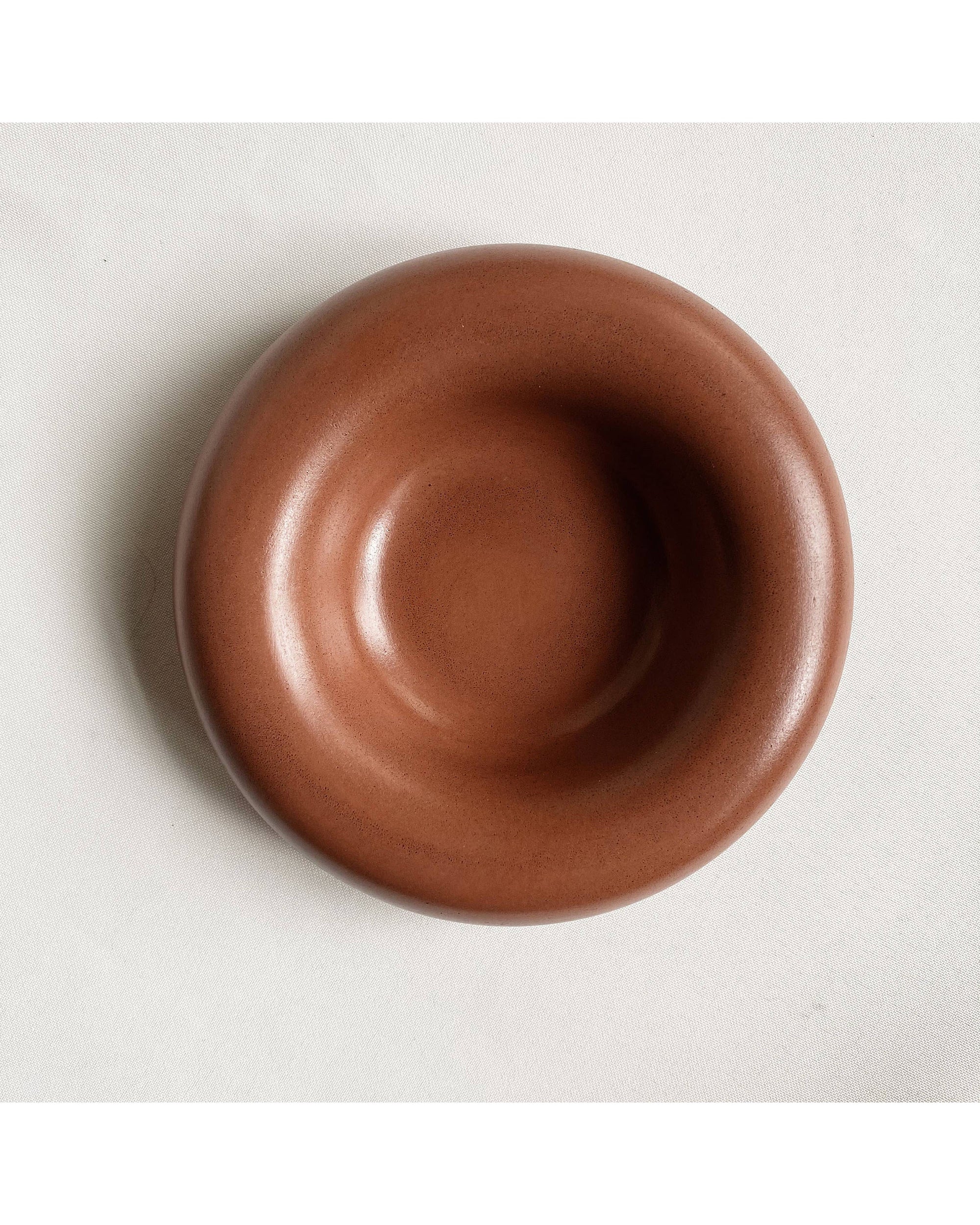 The Faire Gyrus Pocket Tray—a round, smooth, brown ceramic bowl with a wide rim—rests on white fabric atop a concrete tray, seen from above.