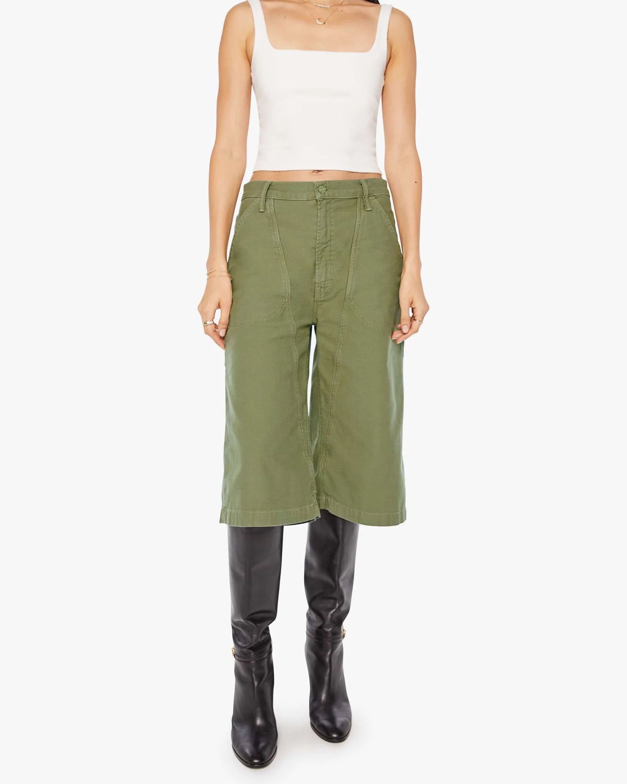 A woman models The Half Chisel by Mother: a white sleeveless crop top paired with army green high-waisted capris and black knee-high boots, set against a plain white background.