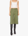A woman models The Half Chisel by Mother: a white sleeveless crop top paired with army green high-waisted capris and black knee-high boots, set against a plain white background.