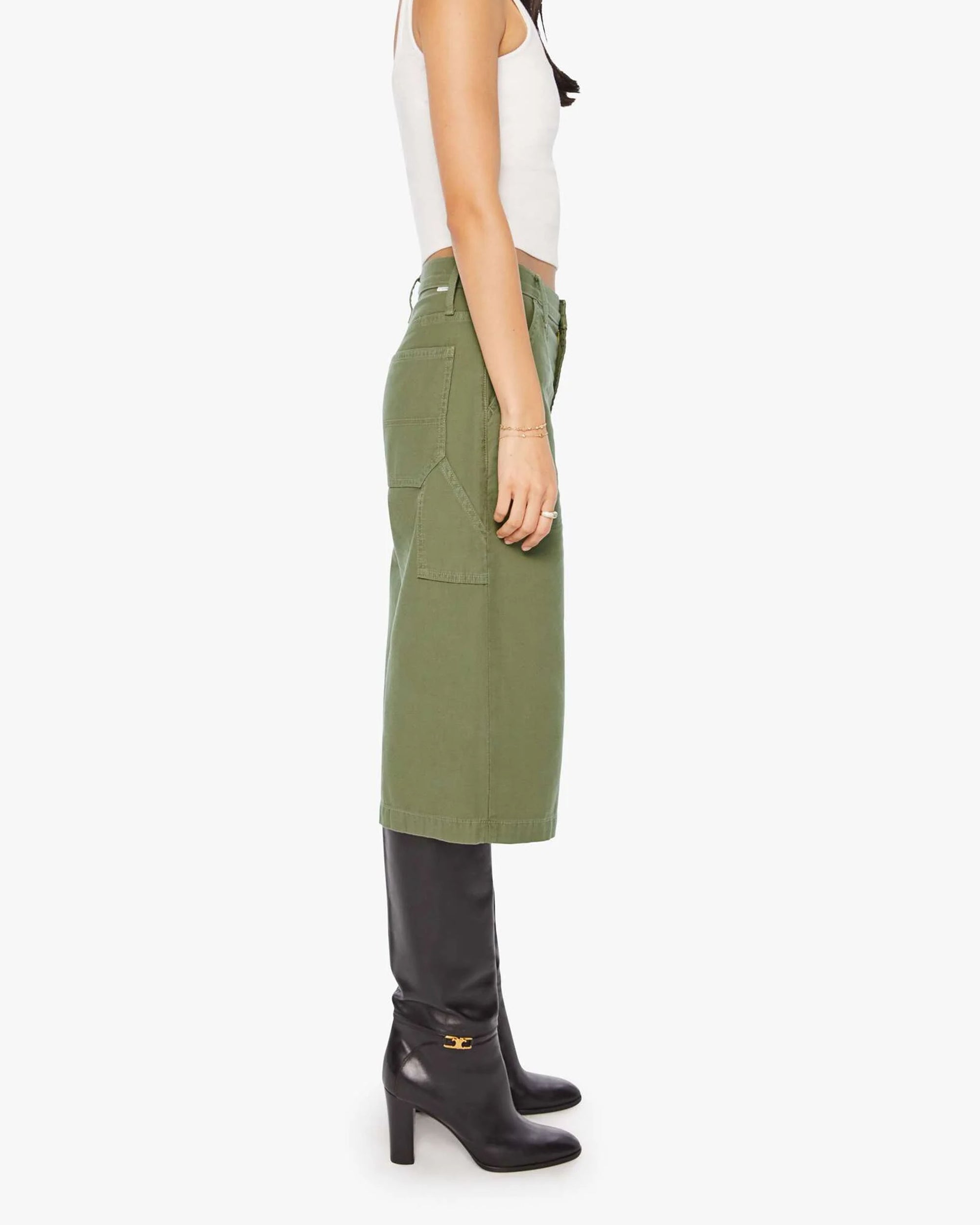 A person stands sideways against a plain white background, wearing Mother’s The Half Chisel: a white sleeveless top and an army green knee-length skirt with utility patch pockets, styled with black high-heeled boots.