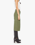 A person stands sideways against a plain white background, wearing Mother’s The Half Chisel: a white sleeveless top and an army green knee-length skirt with utility patch pockets, styled with black high-heeled boots.