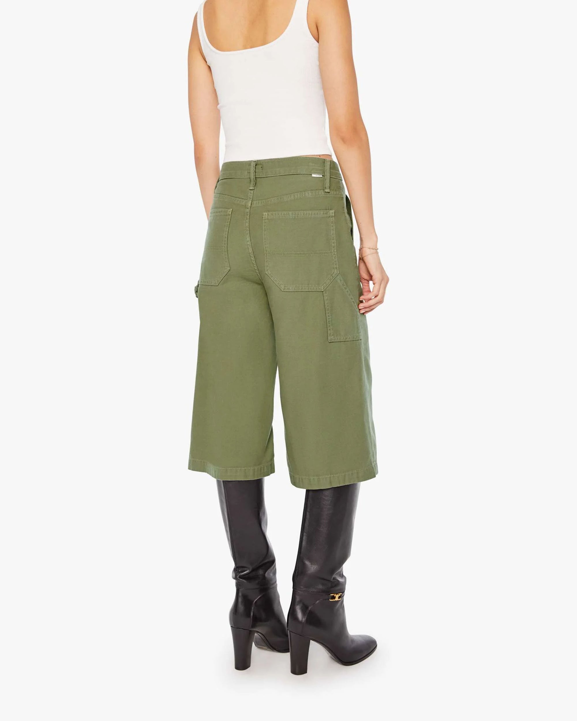 A woman is seen from the back wearing Mother’s The Half Chisel army green high-waisted capris with utility patch pockets, a white tank top, and black heeled boots against a plain white background.