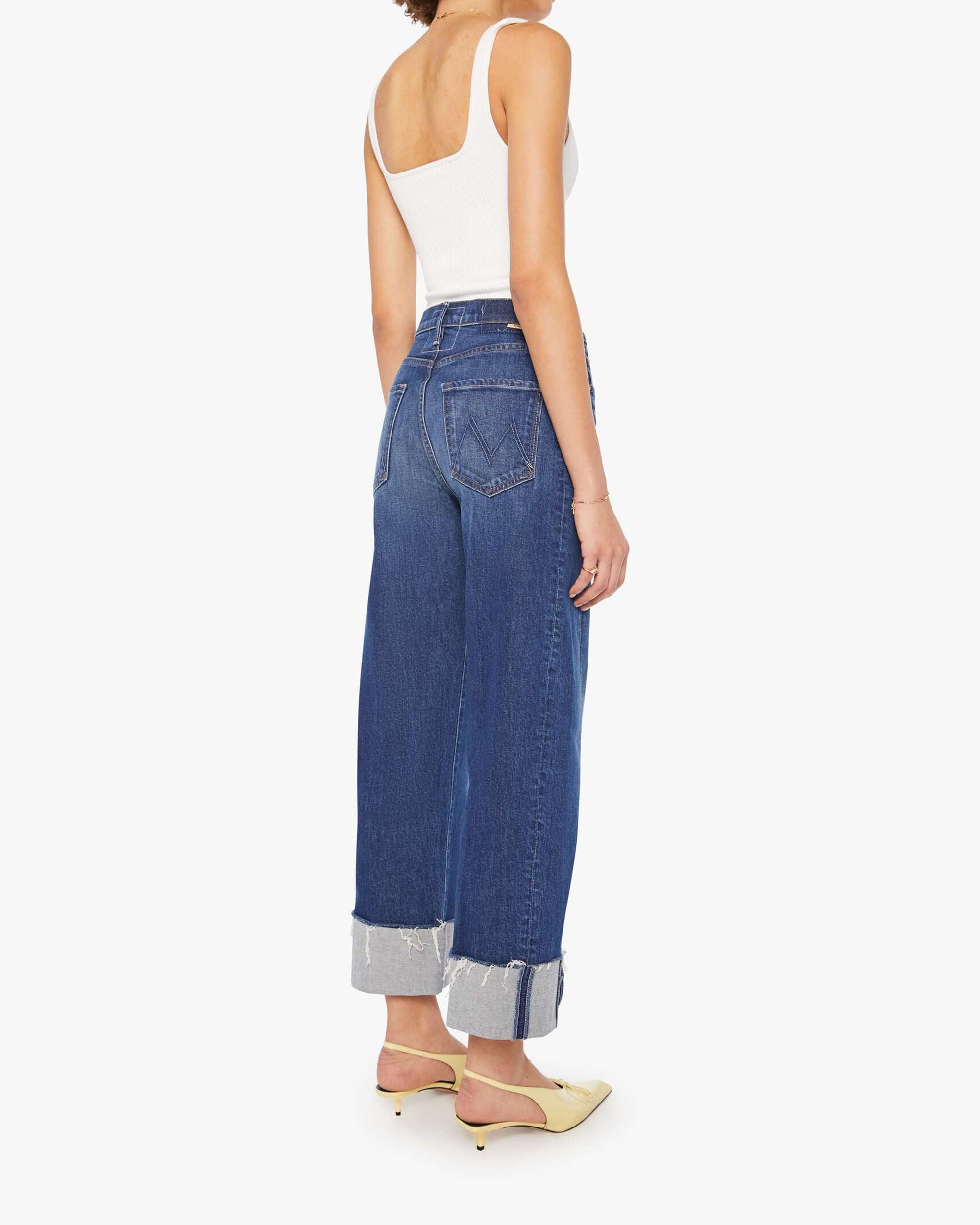 A woman stands facing away, wearing a white tank top, Mother’s Half Pipe Ankle high-waisted barrel jeans in SUPERIOR denim, and light yellow heeled mules against a white background.
