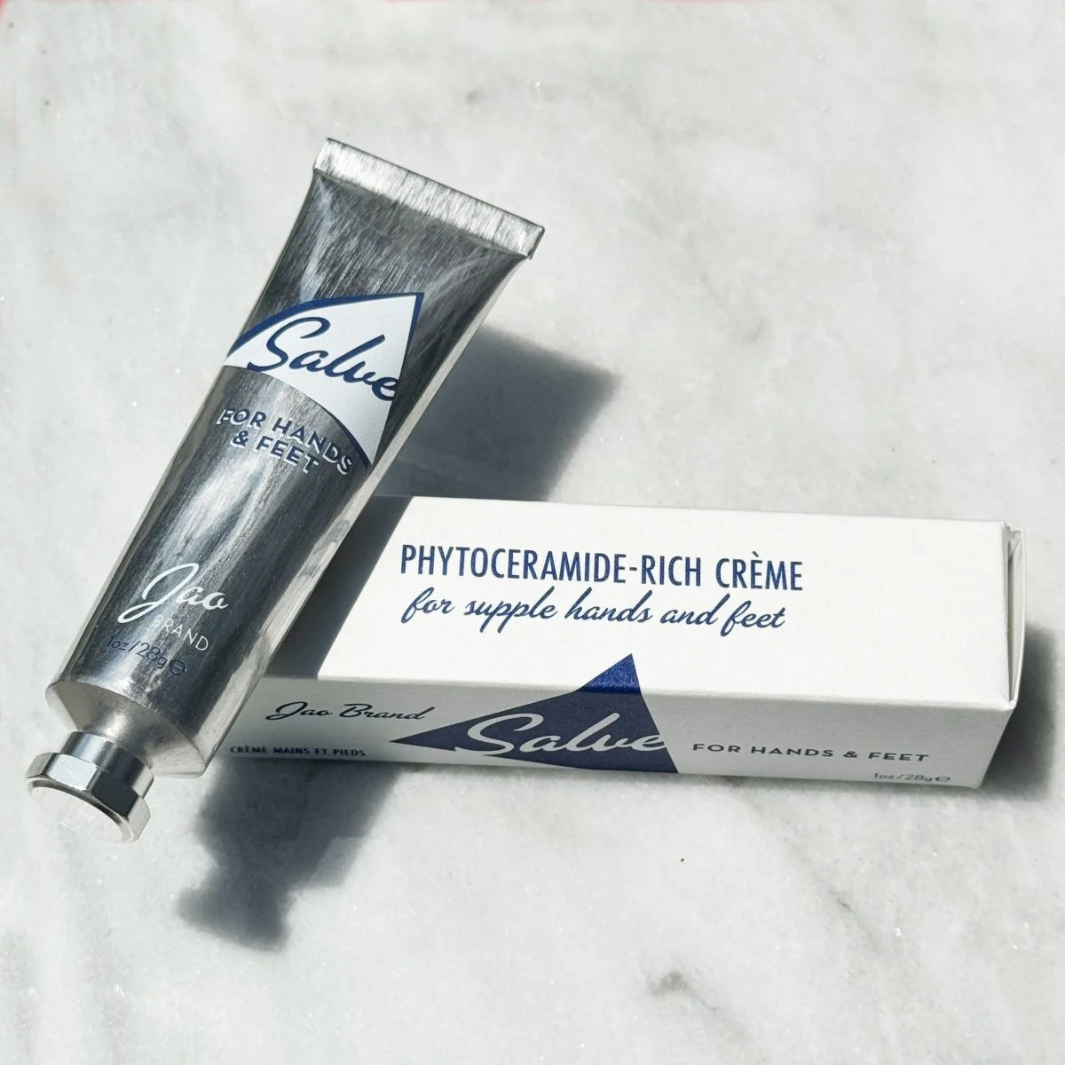 A silver recyclable tube labeled "Jao Hand Salve" by Jao Limited rests next to its white box, which states "Phytoceramide-rich crème to moisturize dry hands and feet," elegantly displayed on a marble surface.