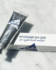 A silver recyclable tube labeled "Jao Hand Salve" by Jao Limited rests next to its white box, which states "Phytoceramide-rich crème to moisturize dry hands and feet," elegantly displayed on a marble surface.