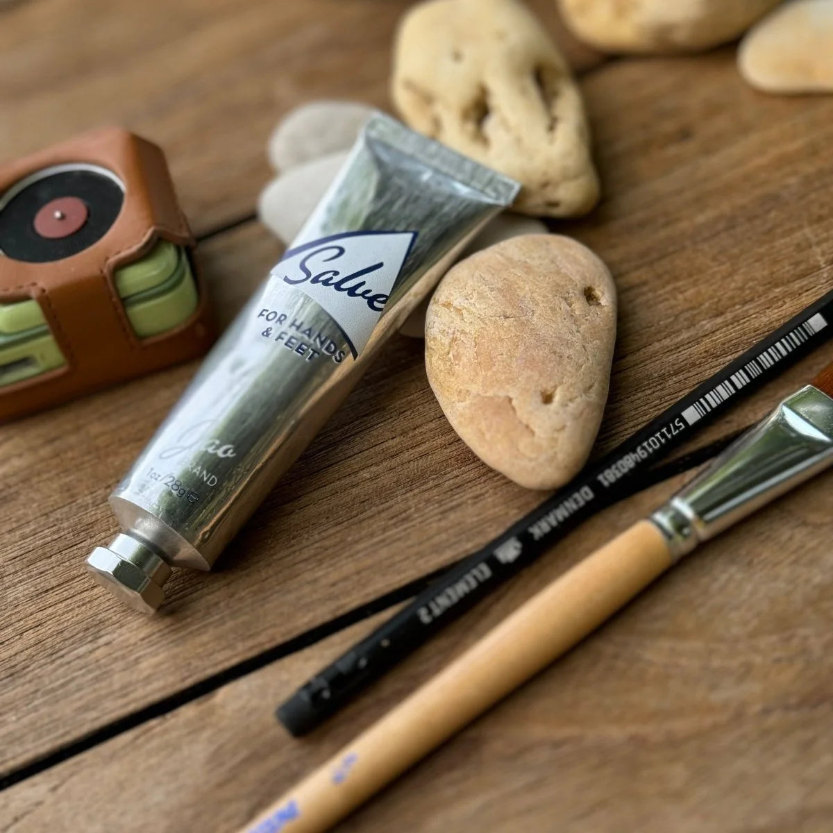 A recyclable tube of Jao Limited’s Jao Hand Salve, a paintbrush, black pencil, smooth stones, and a small brown case with a tape measure are arranged on a wooden surface.