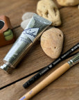 A recyclable tube of Jao Limited’s Jao Hand Salve, a paintbrush, black pencil, smooth stones, and a small brown case with a tape measure are arranged on a wooden surface.