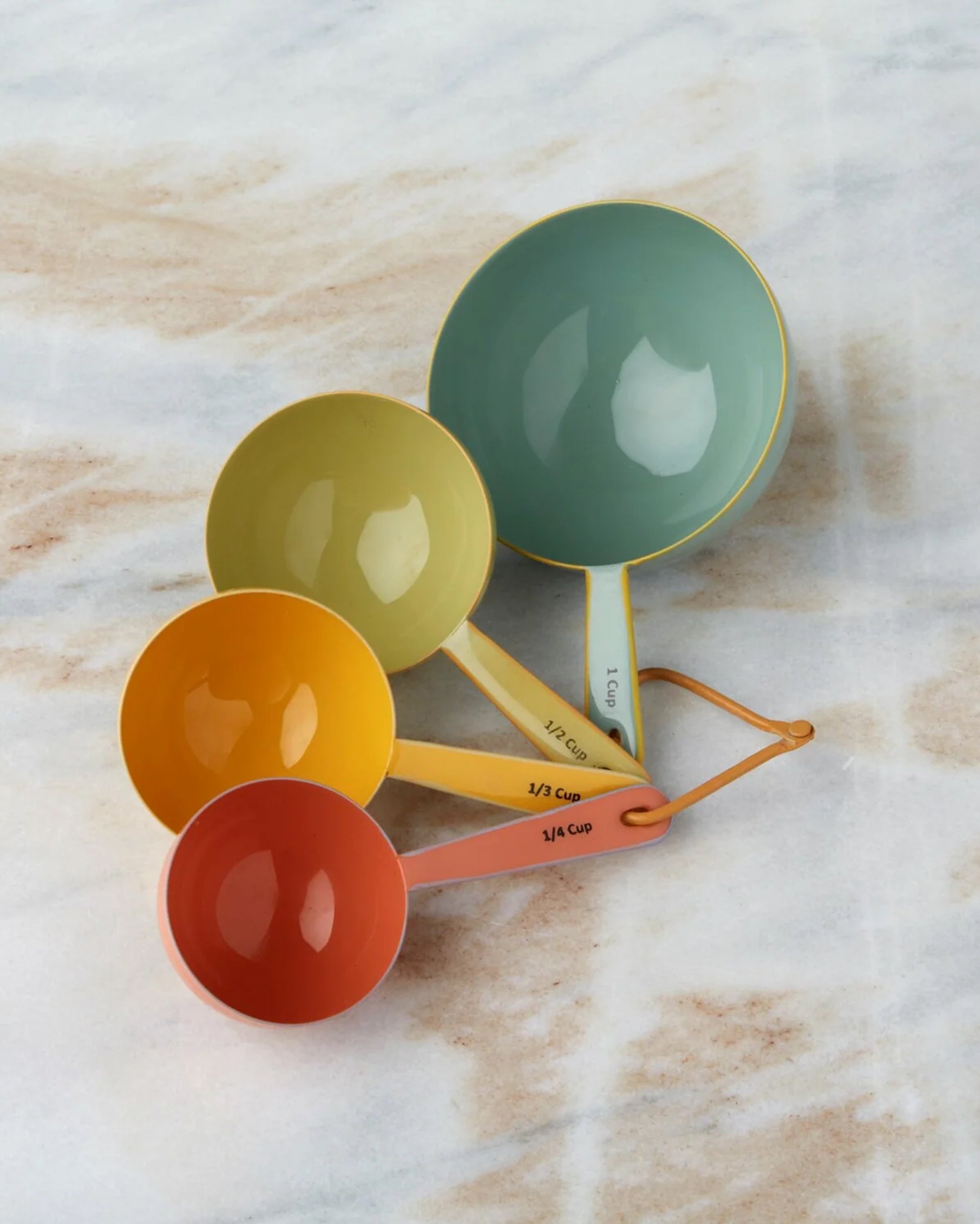 The Harlow Bright Measuring Cups by Be Home feature four enamel-coated cups in red, orange, yellow, and green, each a different size. They're arranged in a fan shape on marble and connected by an orange ring.