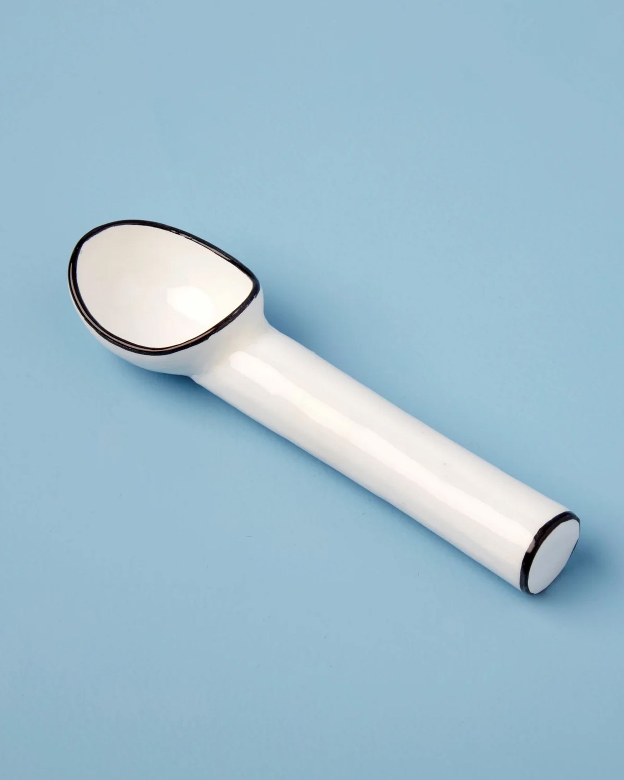 The Harlow Ice Cream Scoop by Be Home, a white ceramic scoop with black-trimmed edges from the Harlow serveware collection, is displayed on a light blue background.