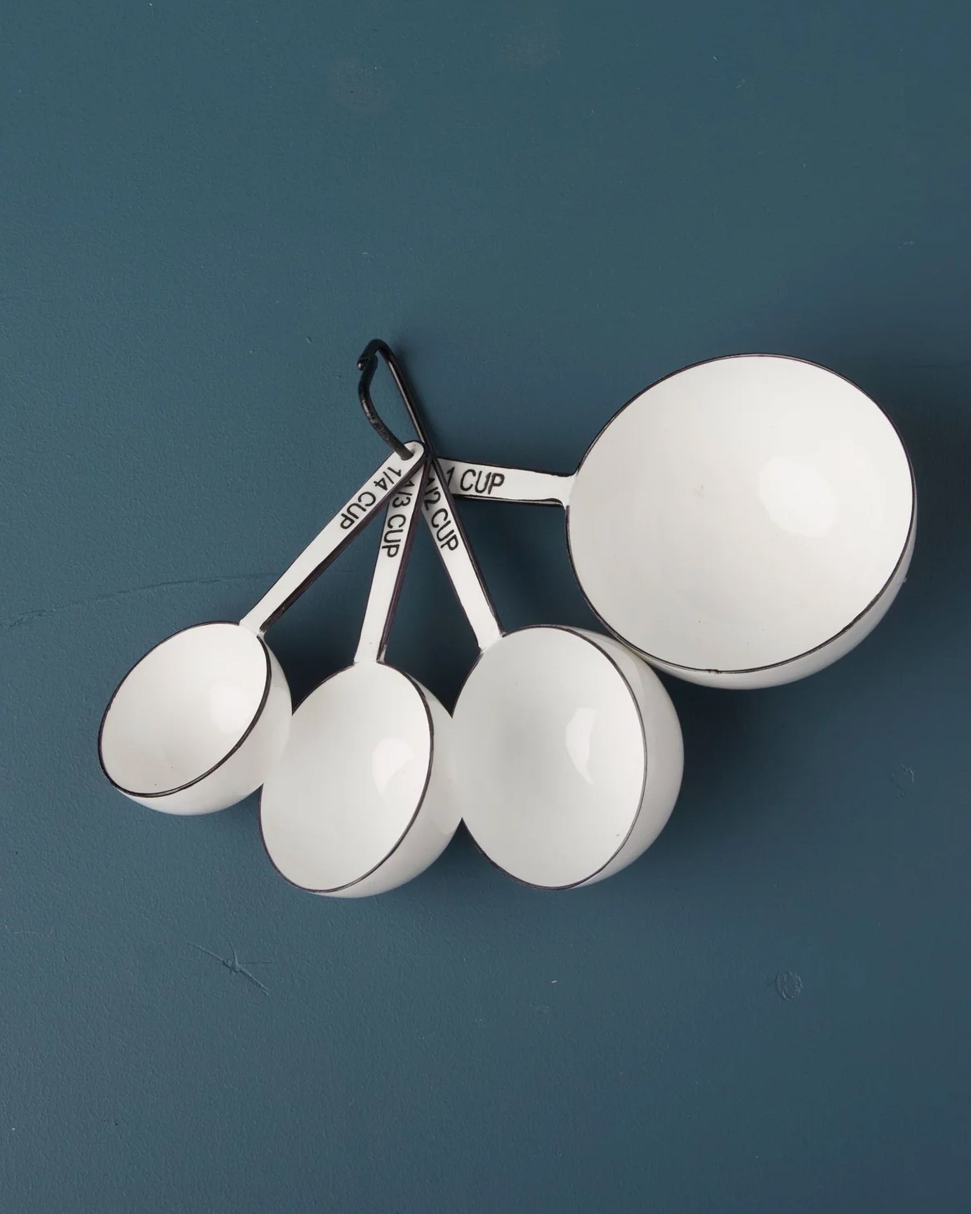 The Be Home Harlow Measuring Cups feature four white cups with black rims and stainless steel handles, labeled 1 cup, 1/2 cup, 1/3 cup, and 1/4 cup. Arranged in a fan shape, they blend vintage style with practical design.