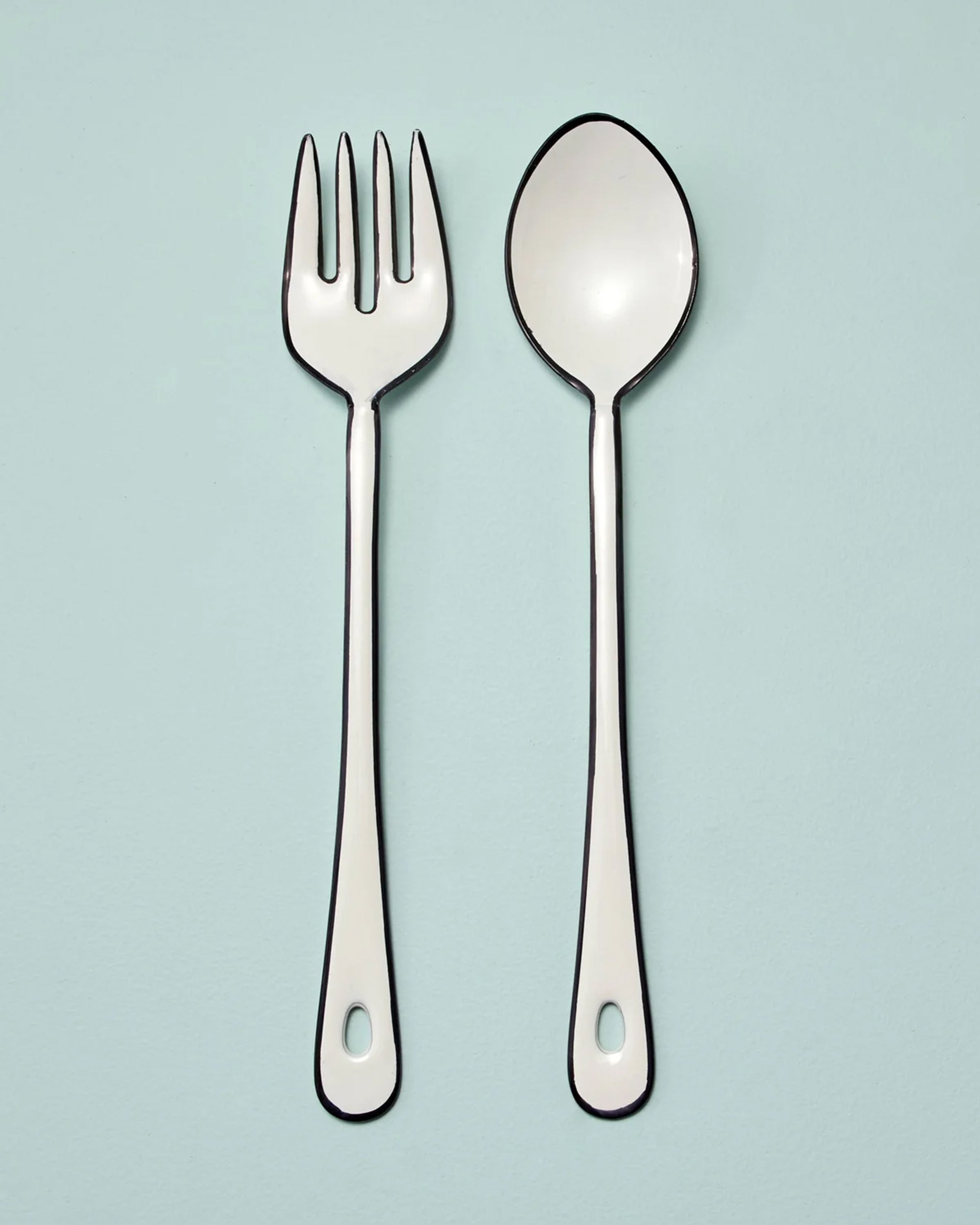 The Harlow Serving Set by Be Home features a white fork and spoon with black outlines and handle holes, placed side by side on a light blue background.