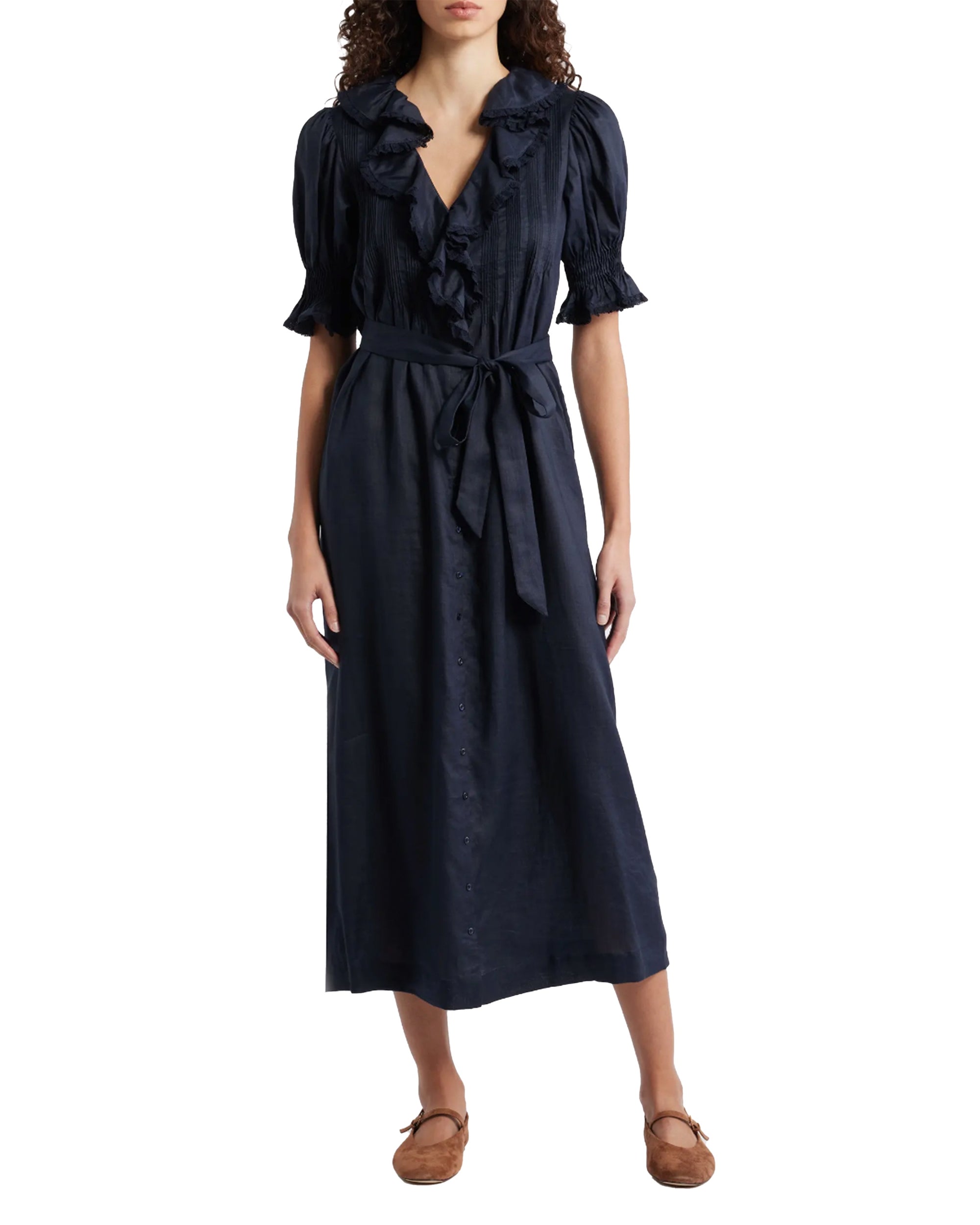 A woman wears the DOEN Harriette Dress—a navy blue midi with short puff sleeves, ruffled V-neckline, button front, lace trim, tied waist belt—and pairs it with brown flat shoes.