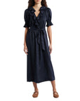 A woman wears the DOEN Harriette Dress—a navy blue midi with short puff sleeves, ruffled V-neckline, button front, lace trim, tied waist belt—and pairs it with brown flat shoes.
