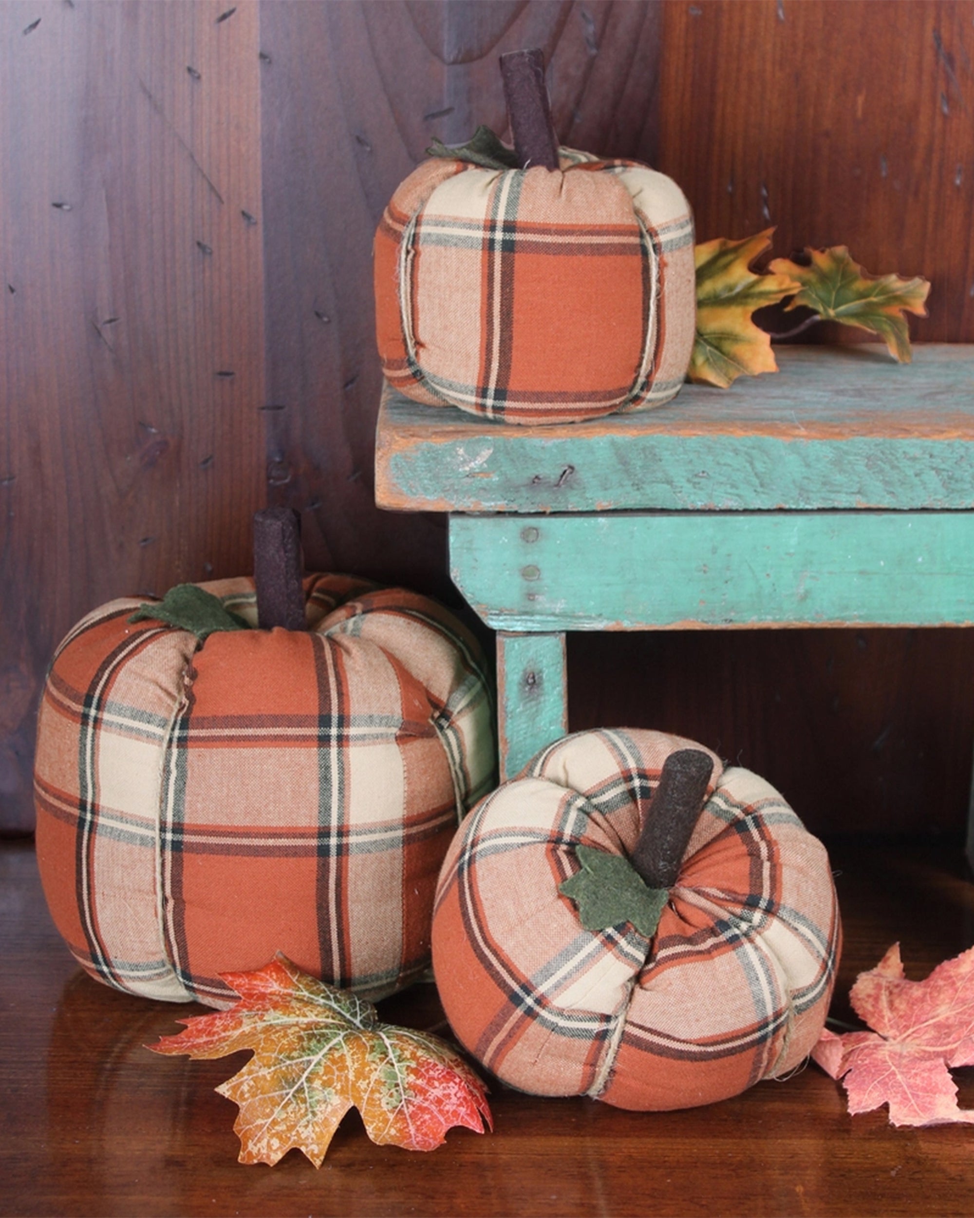 Three Faire Pumpkin decor pieces with orange and cream plaid patterns are arranged among autumn leaves beside a distressed turquoise bench, creating rustic fall charm against a wooden backdrop.
