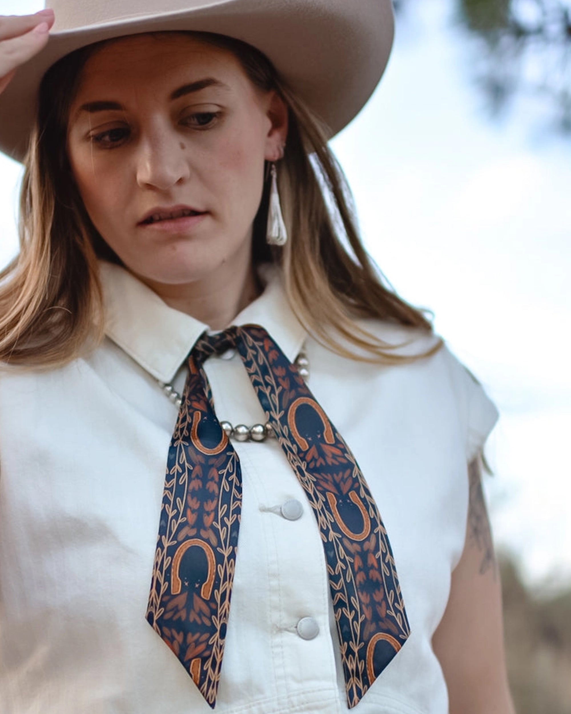 Wearing a wide-brim hat, cream sleeveless button-up shirt, Faire Western Harvest Twilly scarf as a necktie, and drop earrings, the person looks down with a neutral expression.
