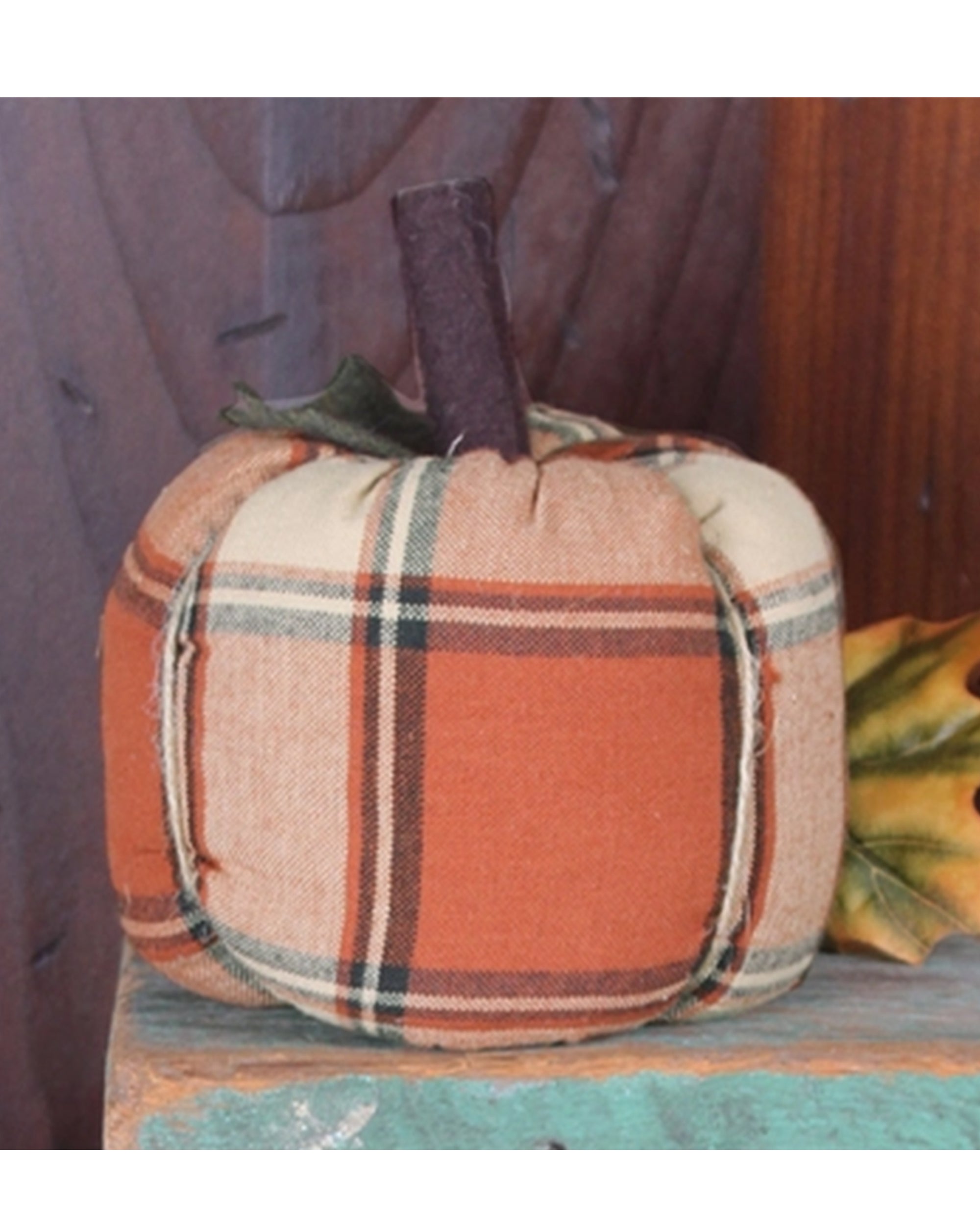 The Faire Pumpkin features a soft orange and tan plaid fabric, ideal for farmhouse decor or fall accents. Displayed on a turquoise wooden surface with a decorative autumn leaf beside it.