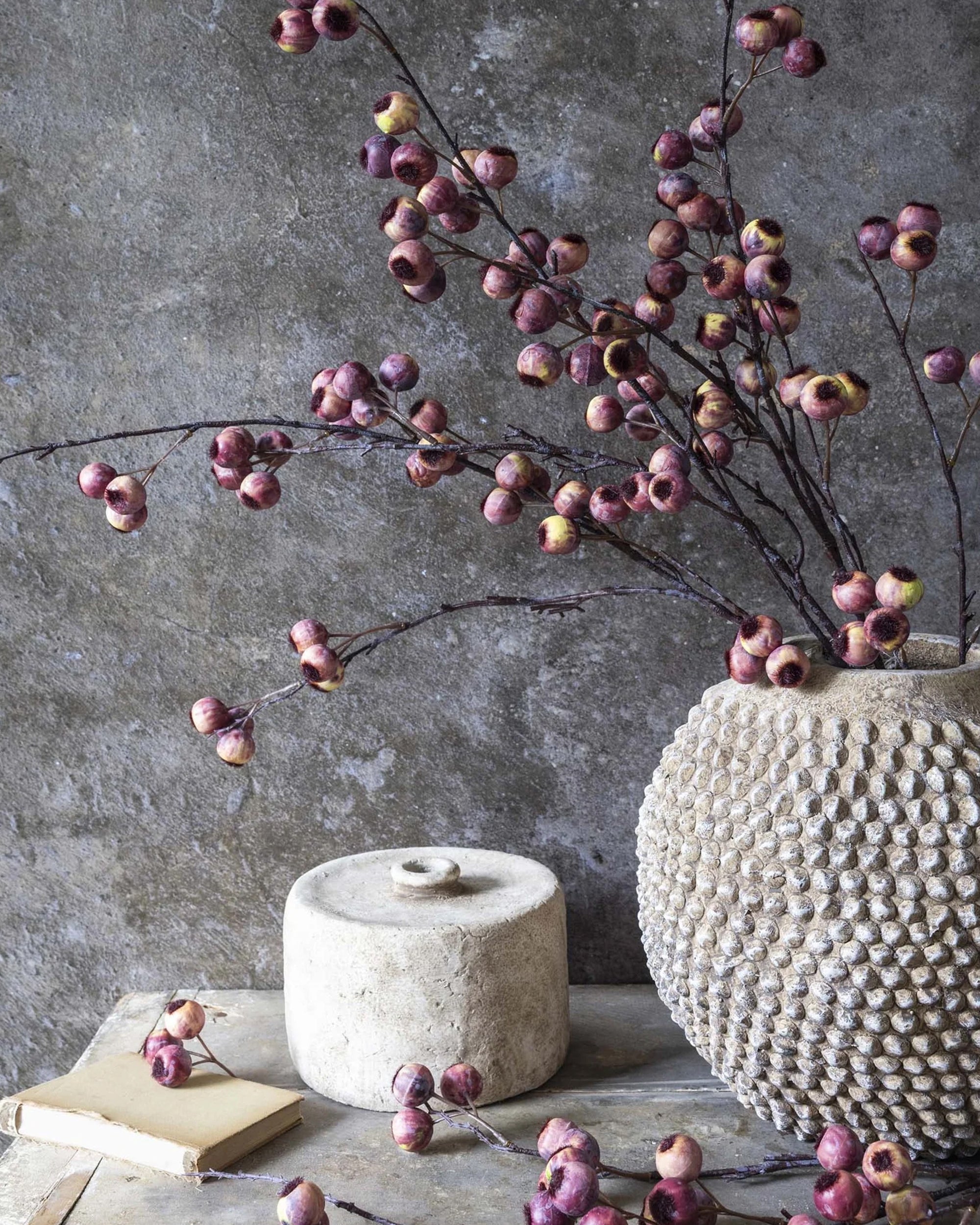 A textured beige Abigail Ahern vase with pink Hawthorne Berry branches sits on a rustic table beside a lidded stone container and a closed book, all set against a rough gray wall.