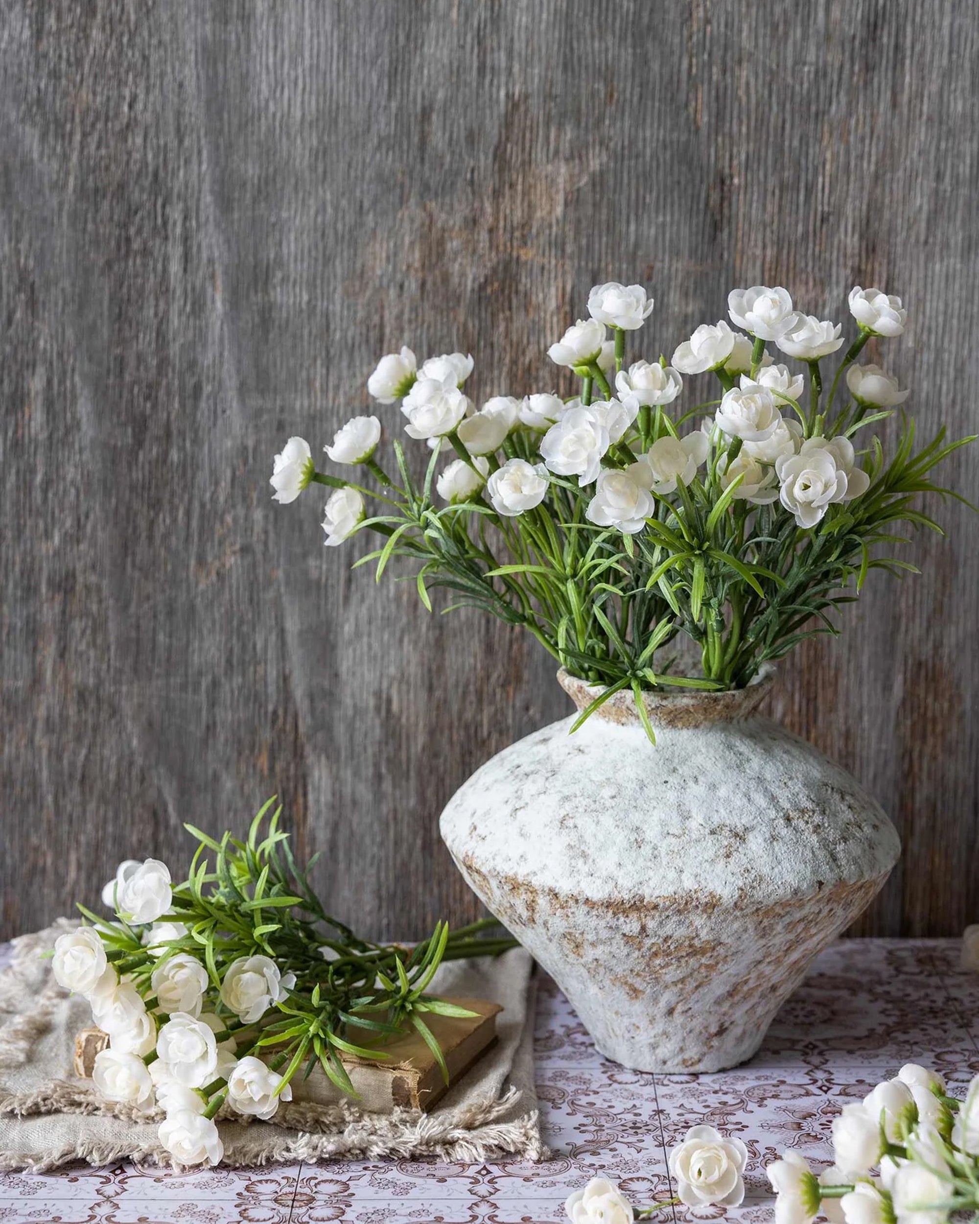 The Hellebore Bunch in White by Abigail Ahern sits in a rustic vase with green foliage; nearby, additional stems rest on lace over a patterned surface, all arranged before a wooden backdrop.