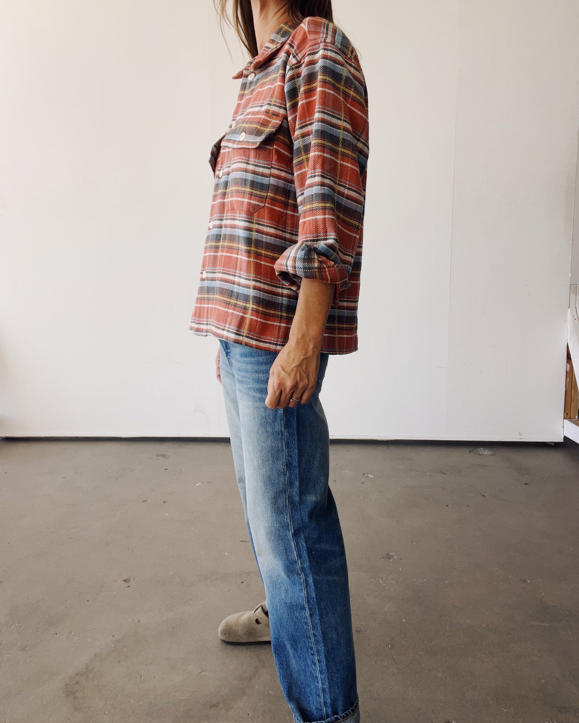 A person stands sideways in a room, wearing Hey Gang’s The Work Shirt in rust plaid with rolled sleeves, paired with blue jeans and gray shoes. The background has white walls and a smooth gray floor.