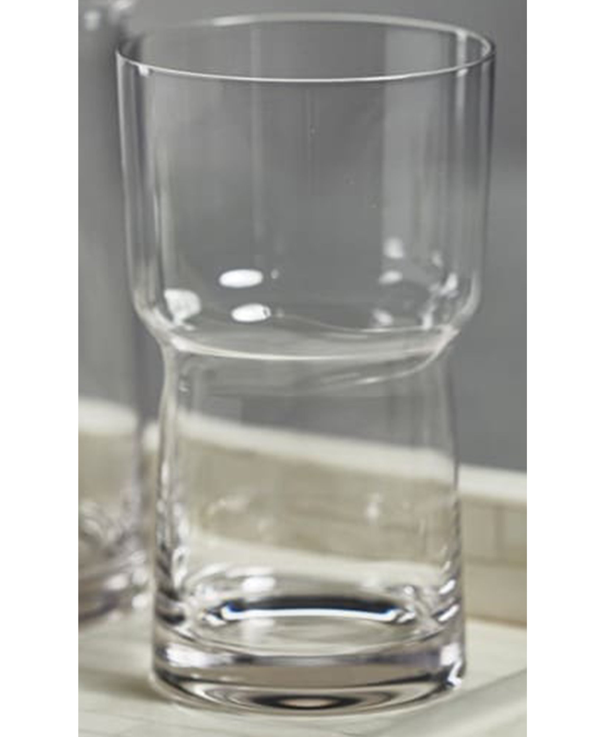 The Zodax Blown Glass HB is a clear, empty highball glass with a unique hourglass shape and thick base, measuring 3.25 x 5.75 inches, displayed on a light-colored surface.