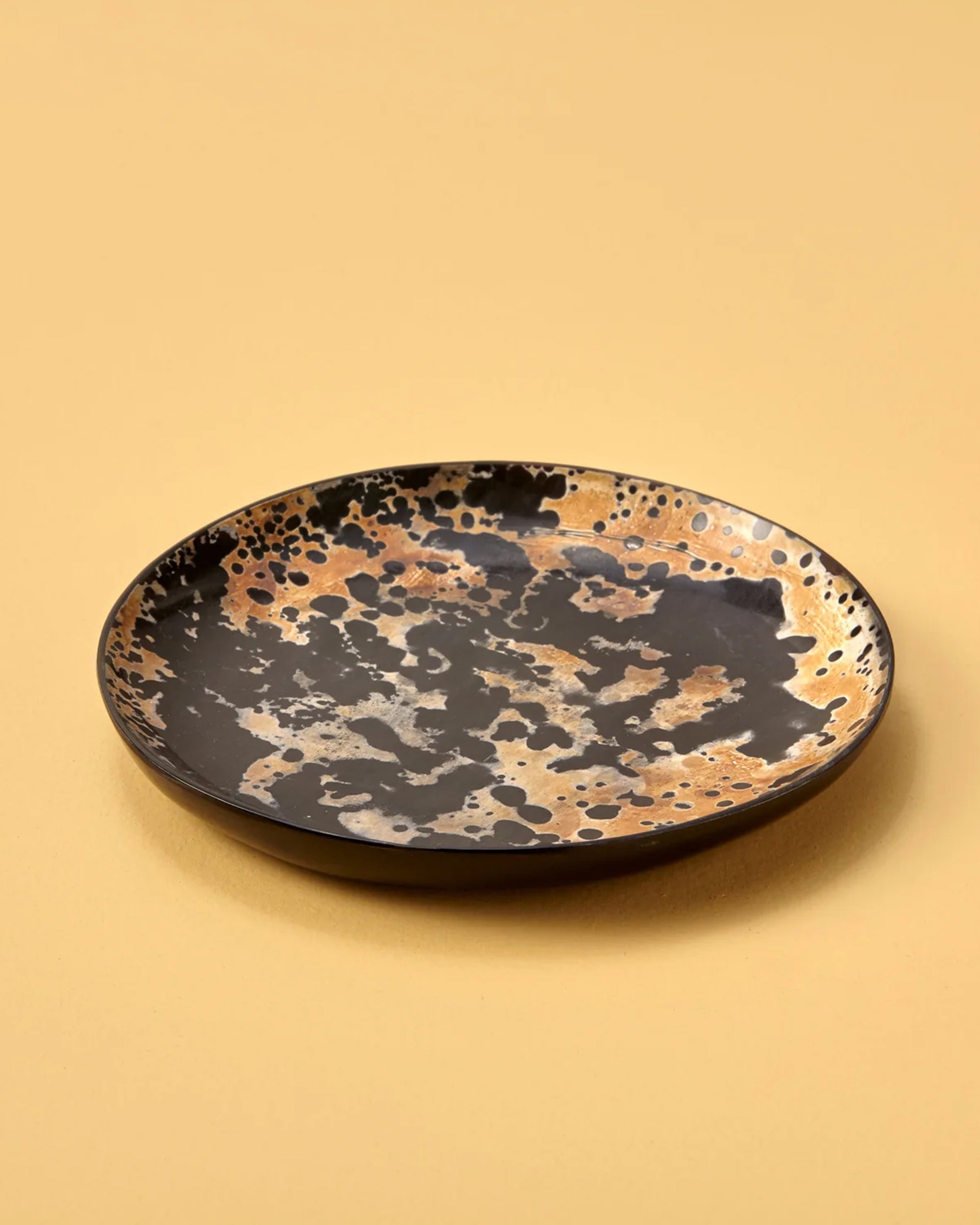 The Be Home Splatter Horn Plate S is a round handcrafted plate featuring a black base with abstract gold and beige splatters, shown on a light tan background.