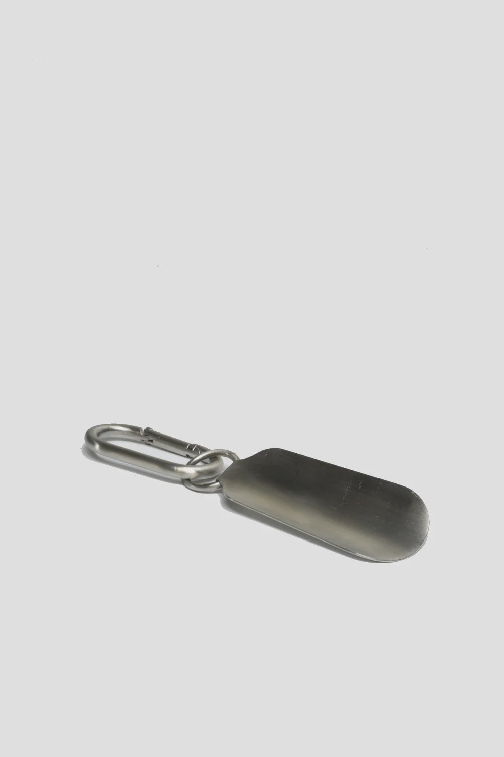 The MENS Steel Shoe Horn by Curated Basics, made entirely from 100% steel and featuring a carabiner clip, stands out on a smooth, light gray background. Its slightly curved design highlights its quality and durability.